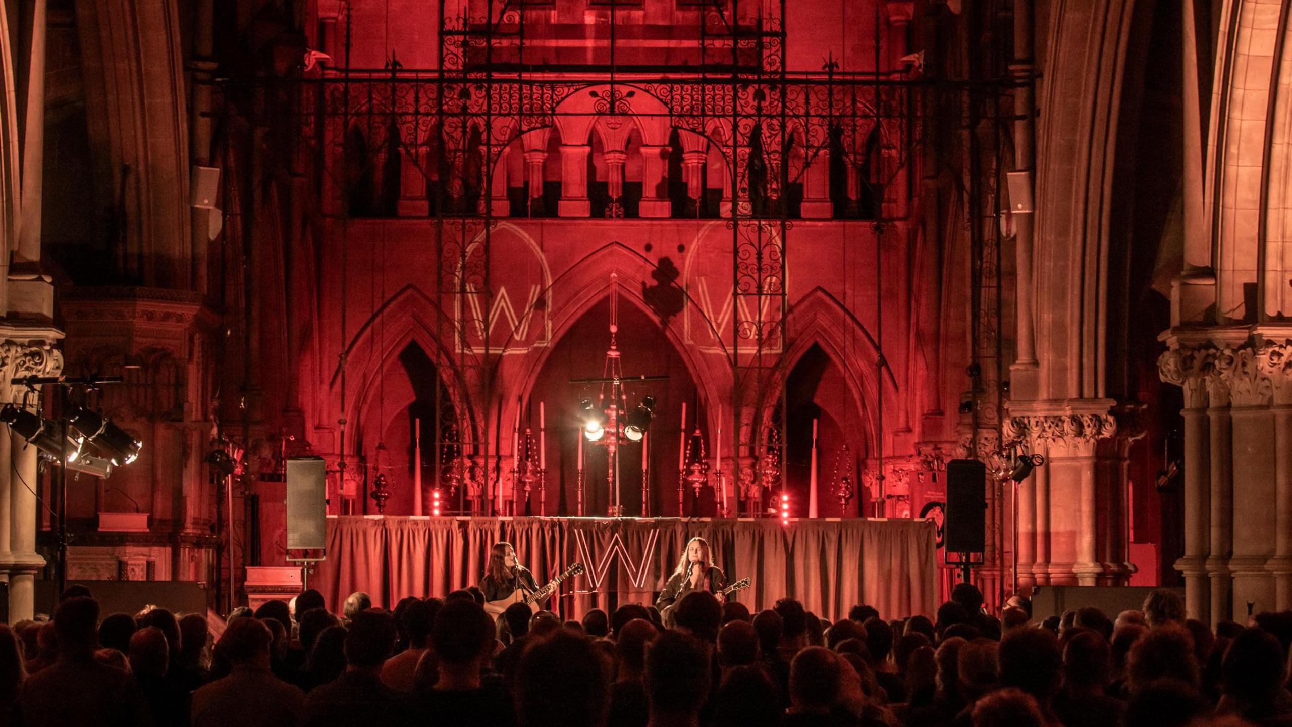 A wide view of The Staves performing at the front of the church with stone arches behind them and a full seated audience in the foreground