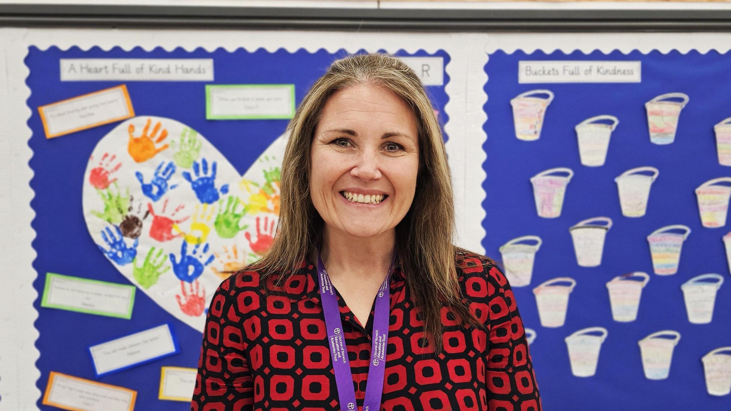 Shannon O'Sullivan is smiling and wearing a black and red patterned top. She's standing in front of displays of childrens' work, including handprints in different colours. 