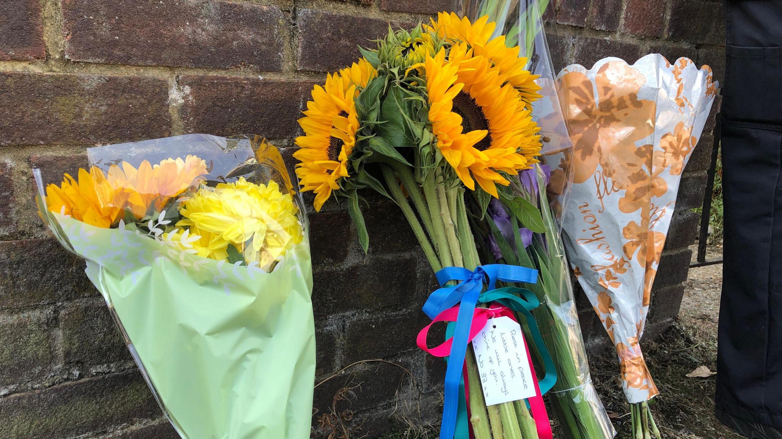 Four bunches of flowers seen on the pavement and propped against a brick wall.