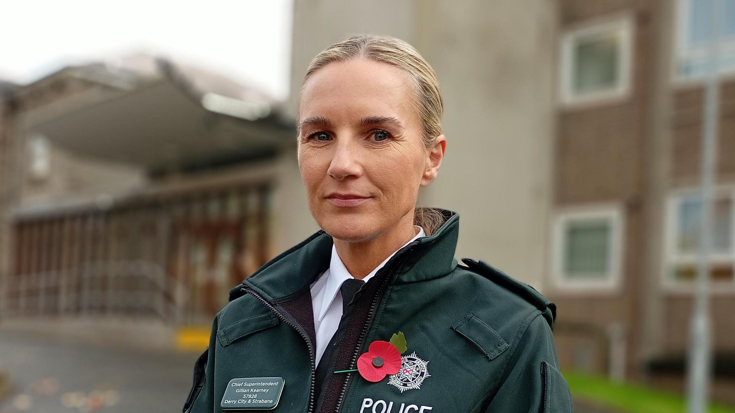 Shows a policewoman in white shirt with hair tied back, while wearing a green jacket. She is also wearing a red poppy on her lapel.