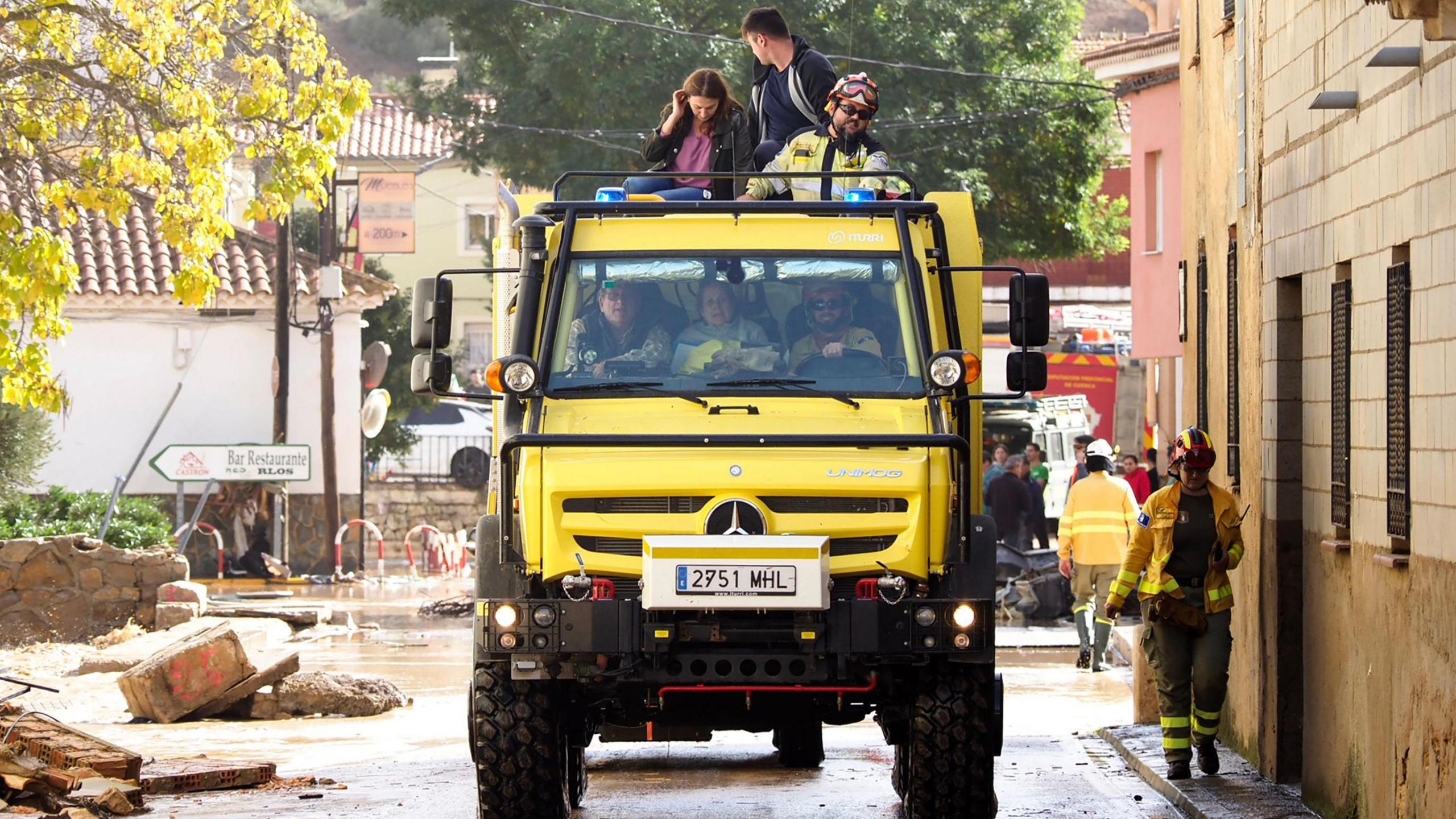 A large yellow truck is driven through a street. There are people inside and on top of the truck. They have been rescued from the flooding. 