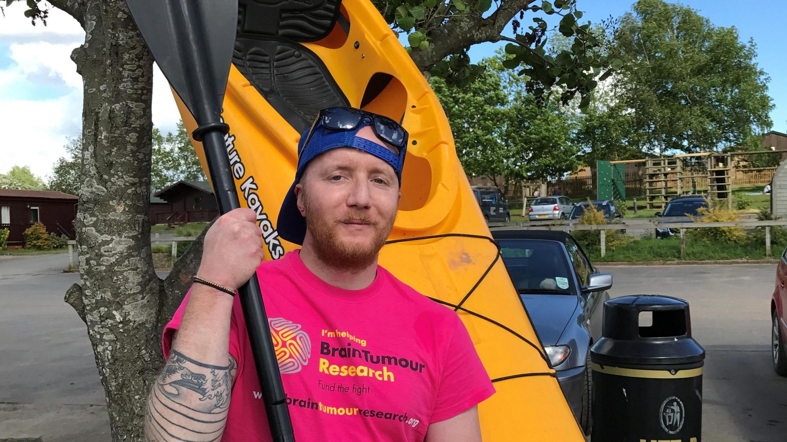 Ben Lindon is leaning against a tree. He is holding a black oar and there is a yellow kayak leaning on the tree behind him. He is wearing a bright pink Brain Tumour Research top, a blue baseball cap and sunglasses on his head. There is a car park behind him with cars parked, a black rubbish bin and some trees.