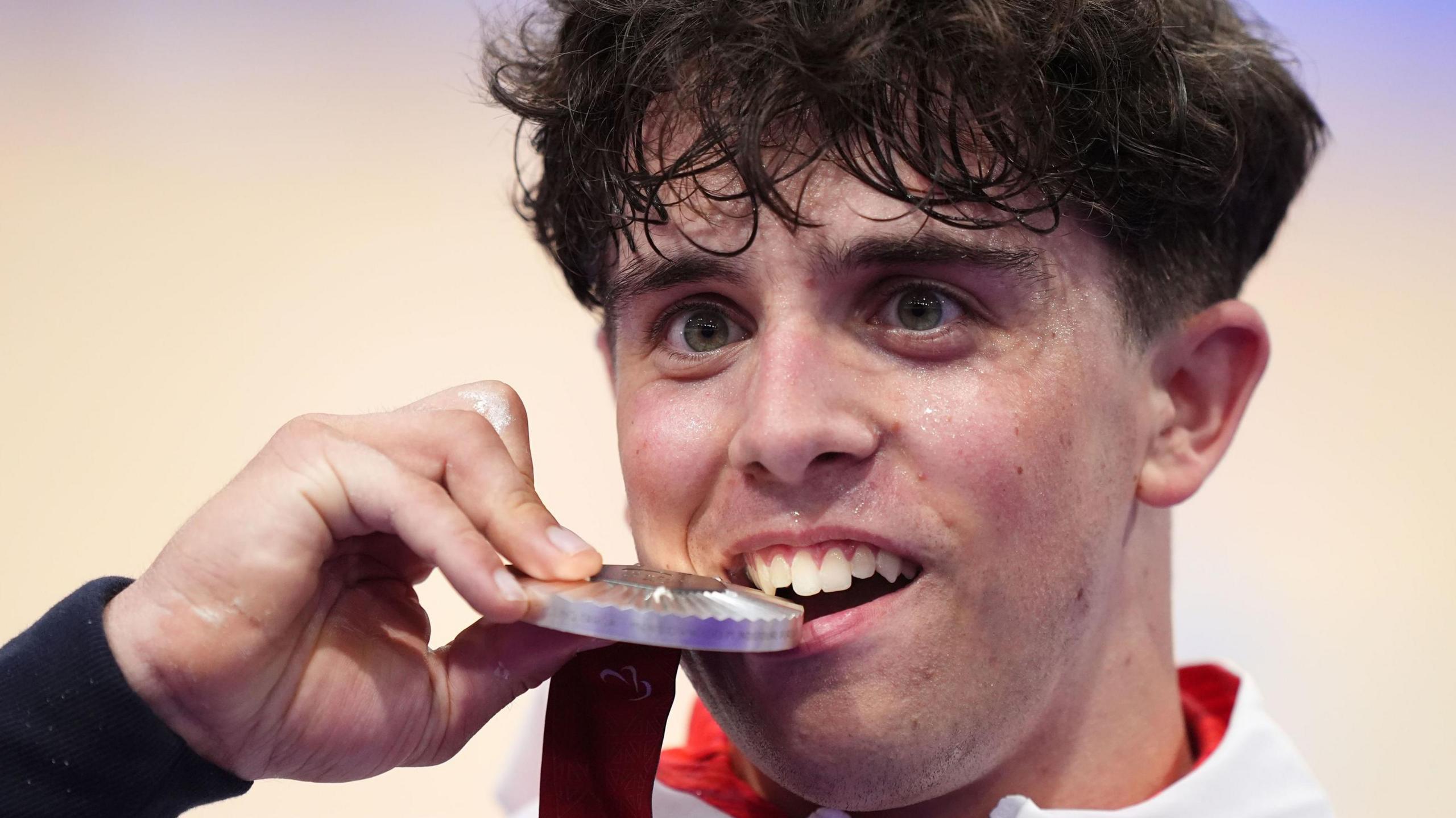 Archie Atkinson at the awards for the Paralympic ceremony for cycling in Paris. He is biting into the silver medal while smiling.