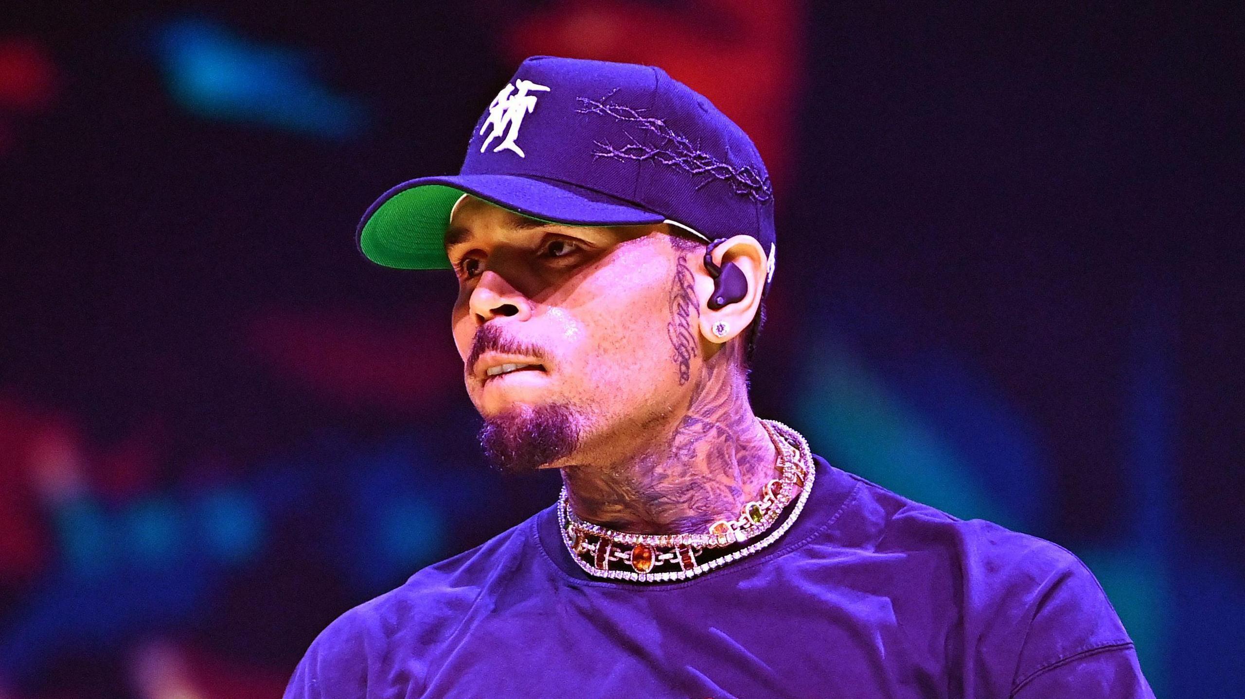 Chris Brown performing in Atlanta in July 2024. He wears a blue baseball cap and matching T-shirt. He has a short goatee beard and tattoos on his neck and the side of his face. 