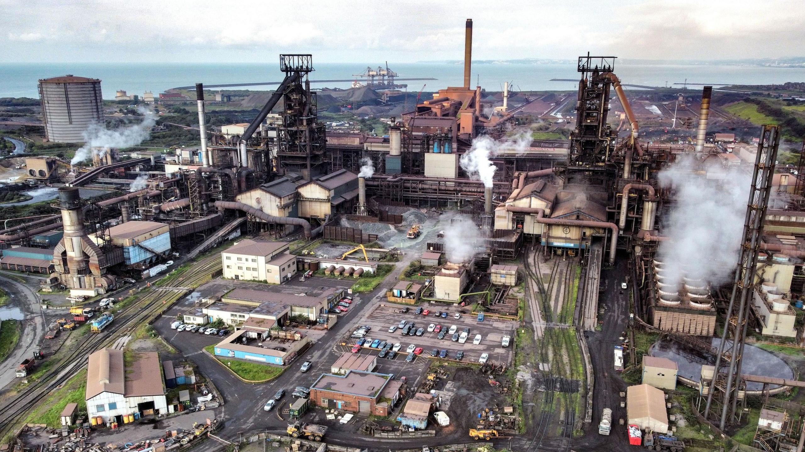 An aerial photo of Tata Steel's Port Talbot steelworks in south Wales.  Smoke and steam is rising from various parts of the plant.  The two blast furnaces are visible along with the sinter plant in the centre of the photo.  