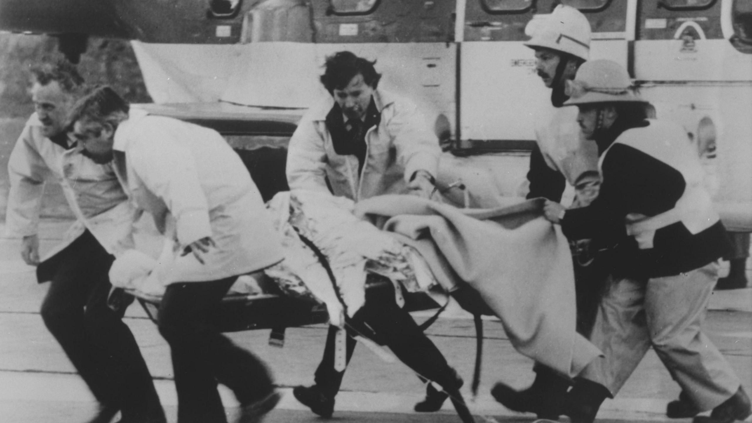 Two fire crew and three medics are rushing a casualty on a trolley from a helicopter. They are moving right to left in frame. In black and white.