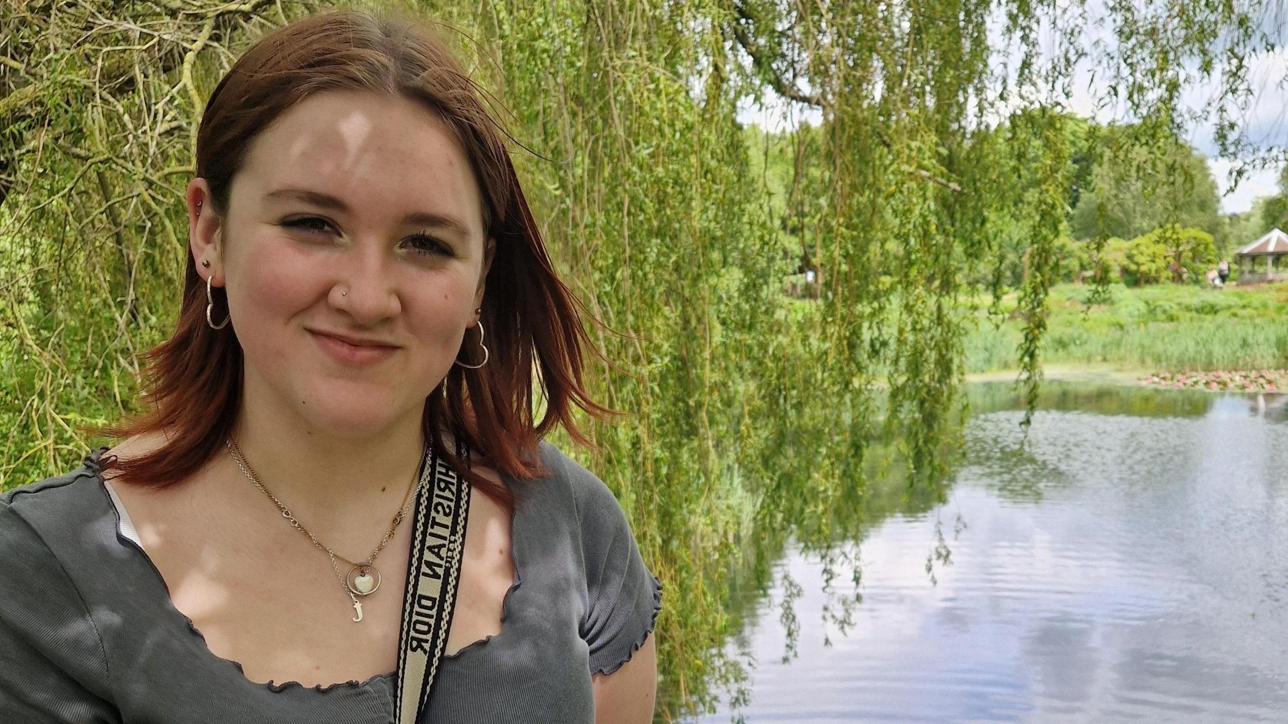 Faye Cullum, wearing a green shirt and a bag strap across her front, stands in front of a lake with tree branches hanging over 