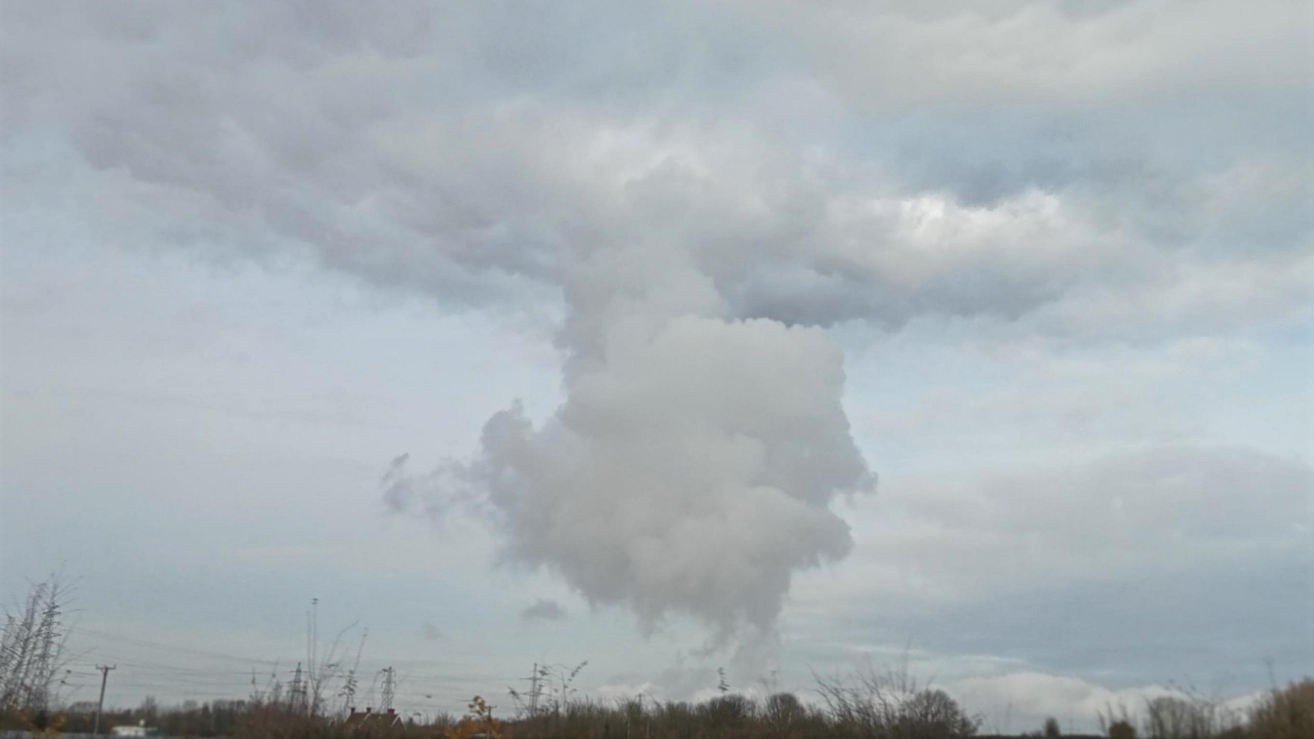 A large plume of grey cloud rises above distant woodland, rising up to a considerable height before flattening out at its top