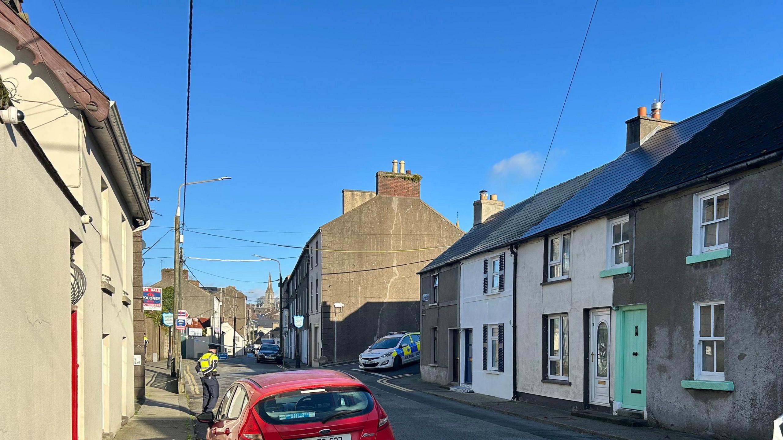 Another view of the street where the investigation is centred.  There are rows of terraced houses on either side of the road.   A garda officer and car are in the background. 