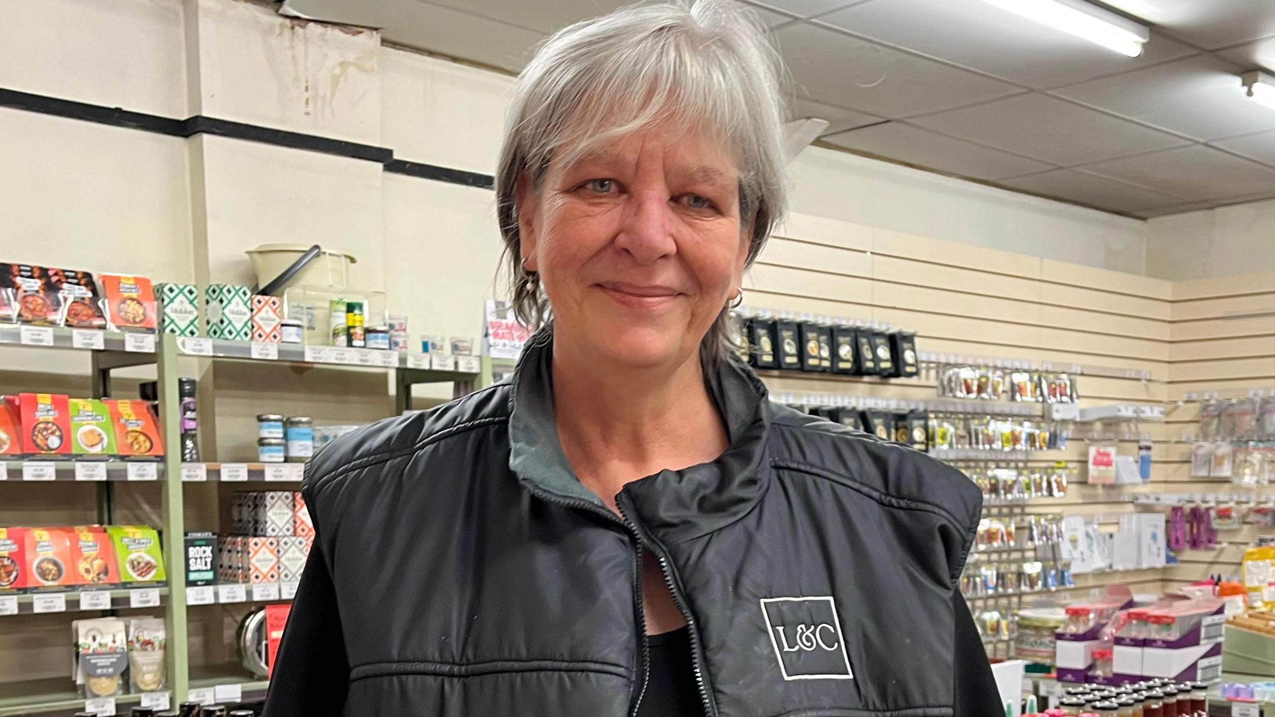 A woman with short grey hair and wearing a black top and black gilet, with an L&C logo on the chest, is standing in front of rows of groceries.