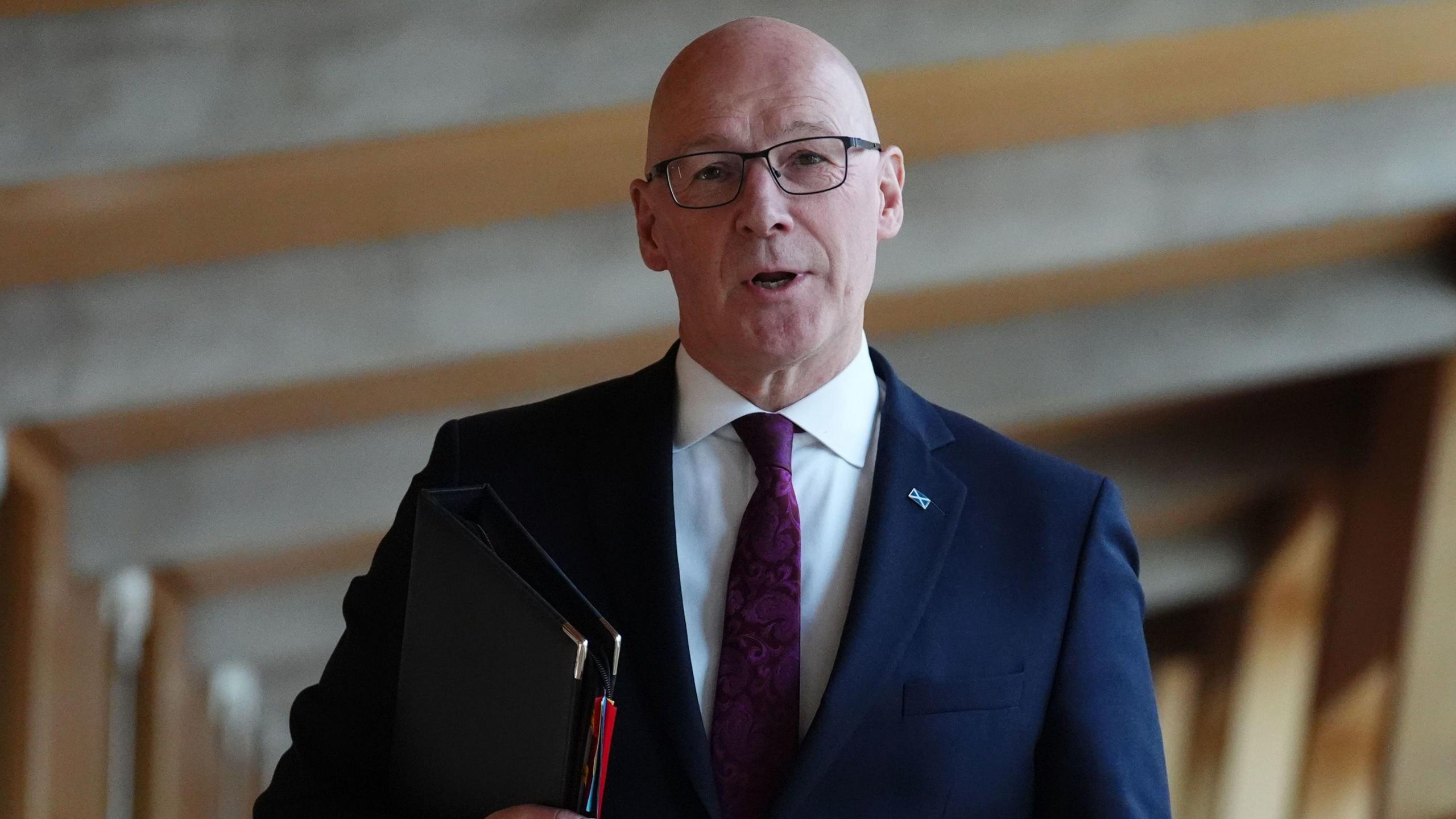 John Swinney in dark blue suit, white shirt and purple tie, carrying a black leather document folder and walking along the long corridor at the Scottish Parliament.