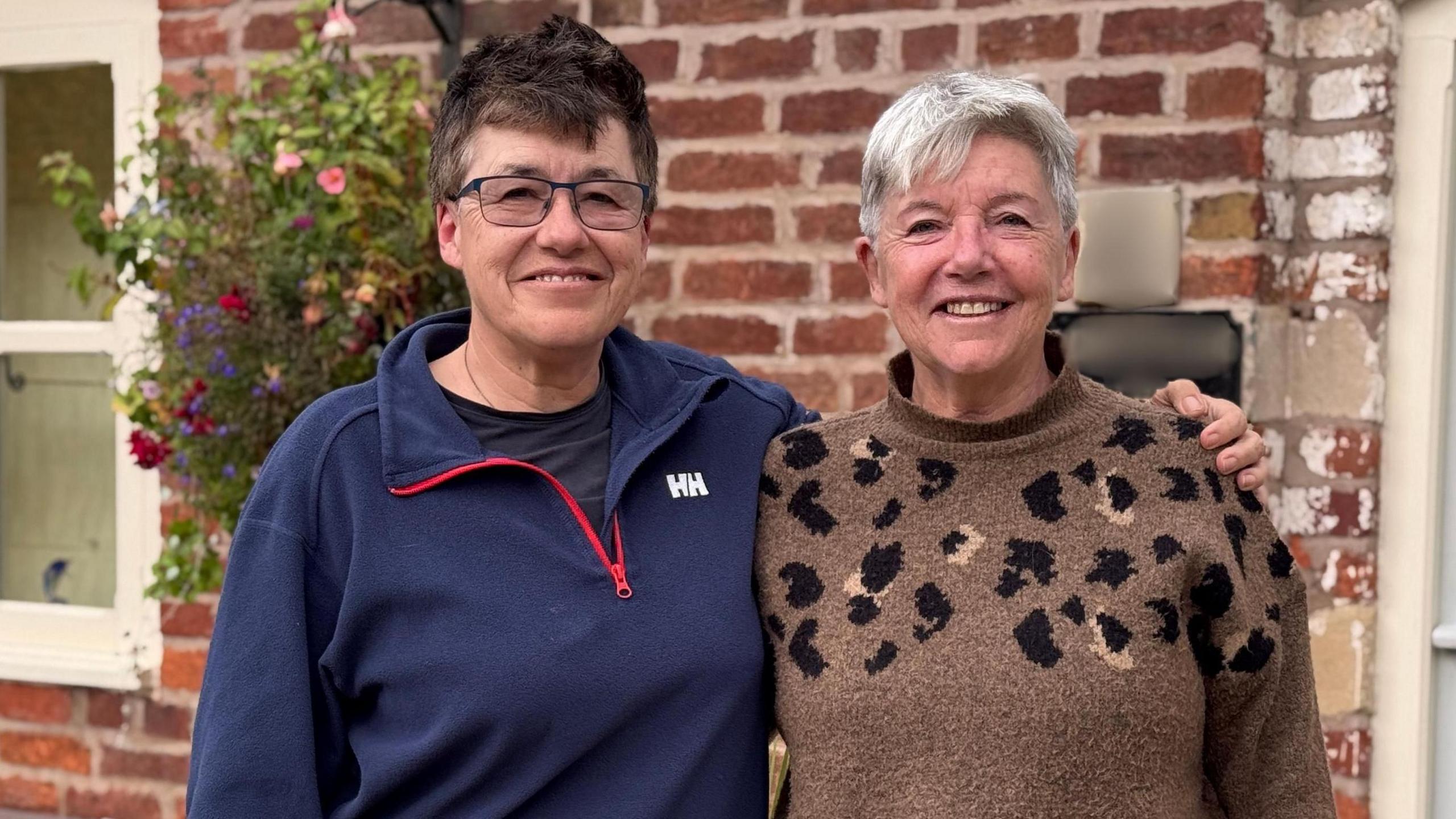Two women are standing next to each other. The one on the left has short dark hair and is wearing glasses and a blue zipped jumper. The woman on the left has short grey hair and is wearing a brown-patterned jumper. They are standing in front of a brick-built house with a grey door. There is a large grey box and flowers nearby.