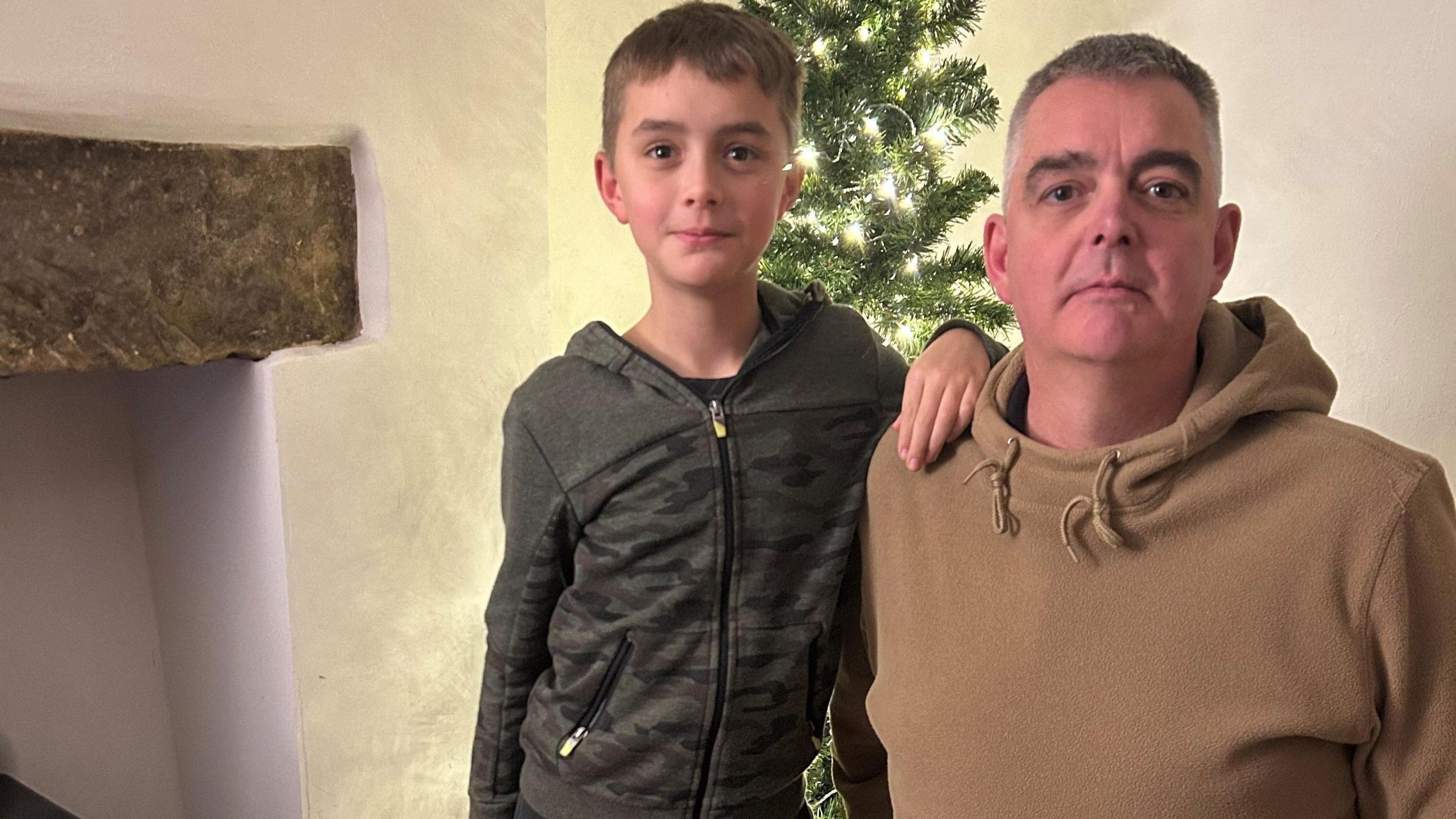 Nick Kirkham crouching next to his son in front of a small Chistmas tree. They're both looking into the camera. Mr Kirkham has got grey hair and is wearing a brown hoodie. 