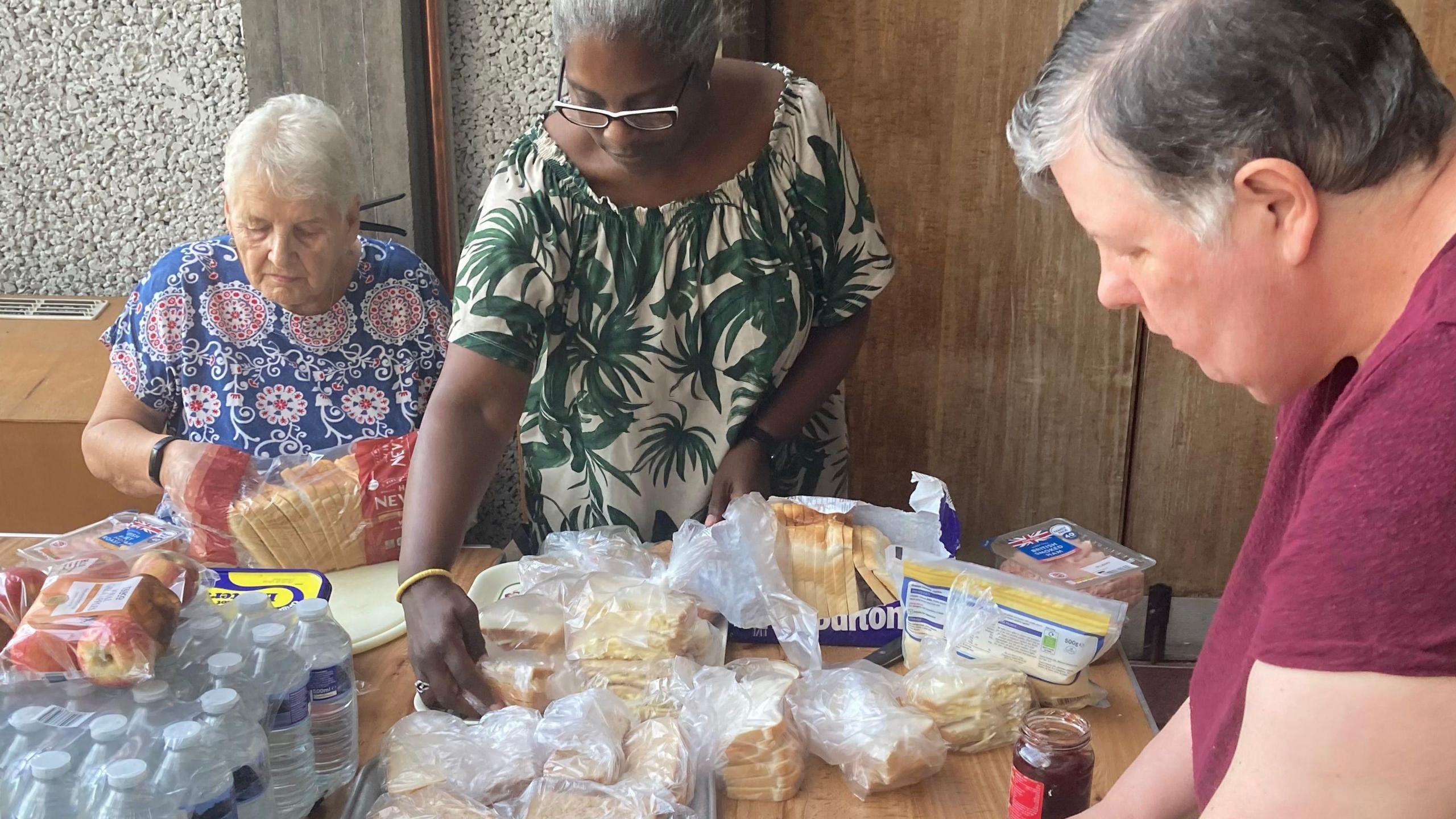 Volunteers are working at a table in a church building making and laying out sandwiches wrapped in clingfilm on a tray. Supplies of bottled water are to one side. One of the volunteers is making the sandwiches and another is wrapping them and stacking them up. The room has a door with fire exit notices on it. The women are wearing summer dresses.