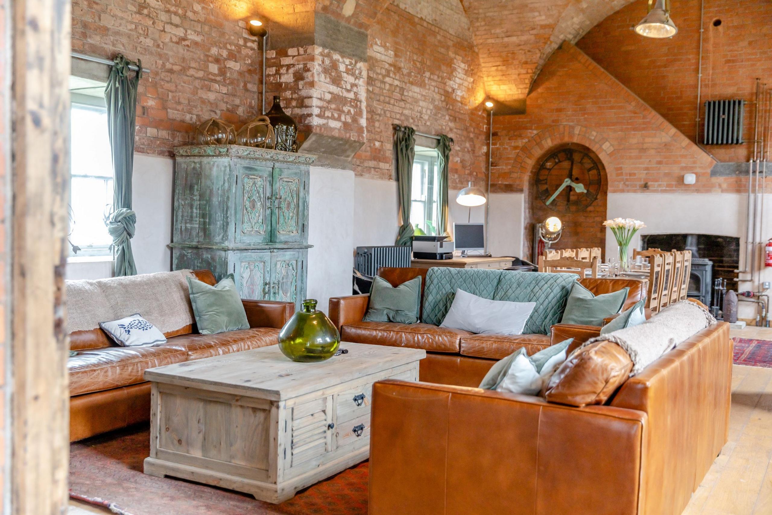 A living room with high ceilings and red brick walls. Three leather sofas face a chunky wooden coffee table, with a dining table behind it. 