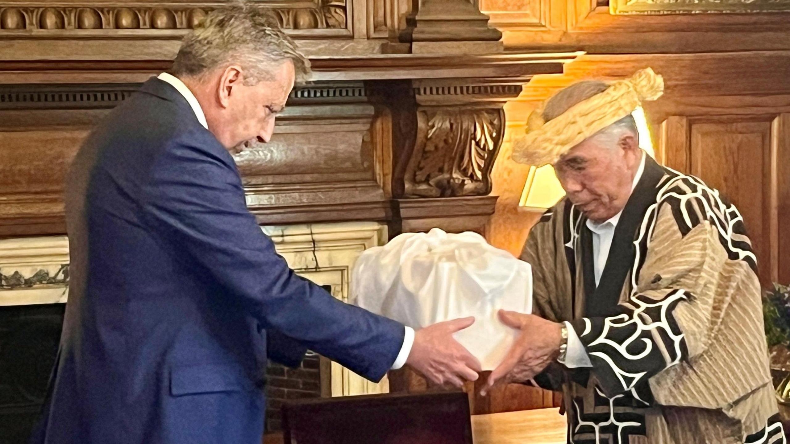 Edinburgh University principal Sir Peter Mathieson, wearing a blue suit, hands a box covered in white cloth to a man wearing a beige and black-coloured robe over a white shirt.