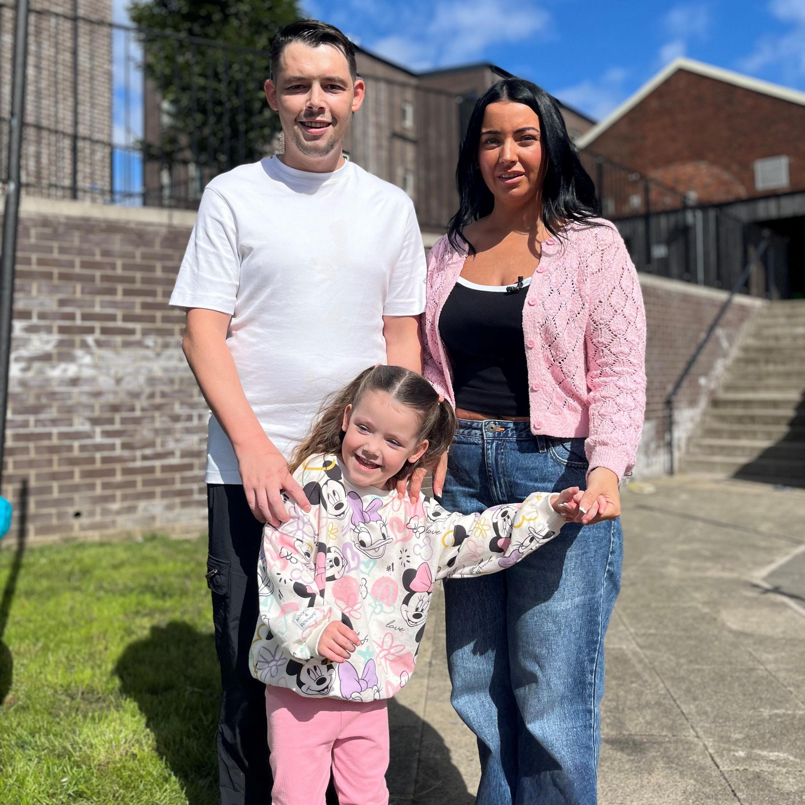 Pic of Niamh with dad Liam and mum Samantha> They are stood outside with a wall and steps in the background. Niamh is wearing a fleece jacket with Mickey Mouse patterns on it and pink trousers. Her mother has long black hair, a pink cardigan and blue jeans. Her dad has short black hair, a white T-shirt and jeans