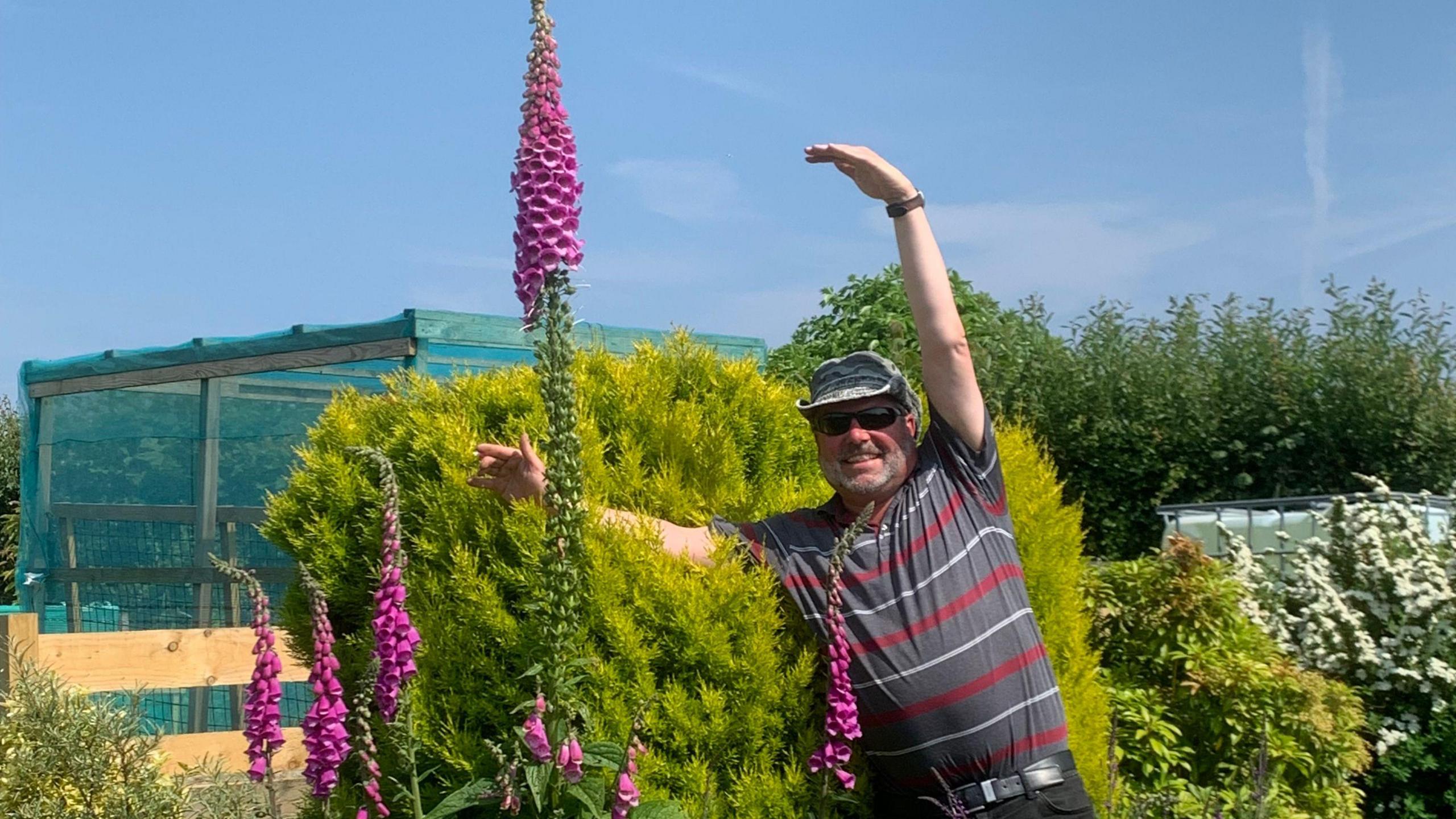 A man with grey stubble and a beige sun shader hat wears a grey t-shirt and poses with his hands in the air in a garden with tall pink flowers, on a sunny day.