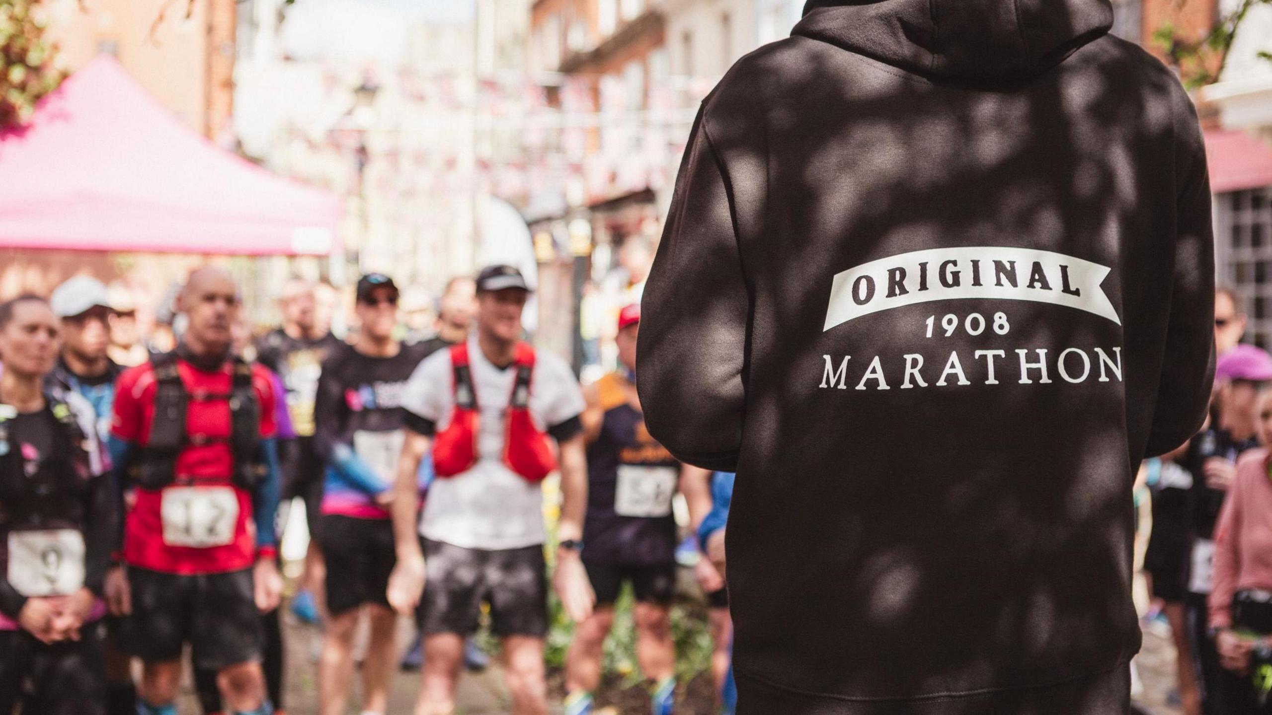 Blurred runners face towards the camera and a person is standing closer to the camera but facing away with the back of their black hooded sweatshirt in shot with Original 1908 marathon logo printed in white.