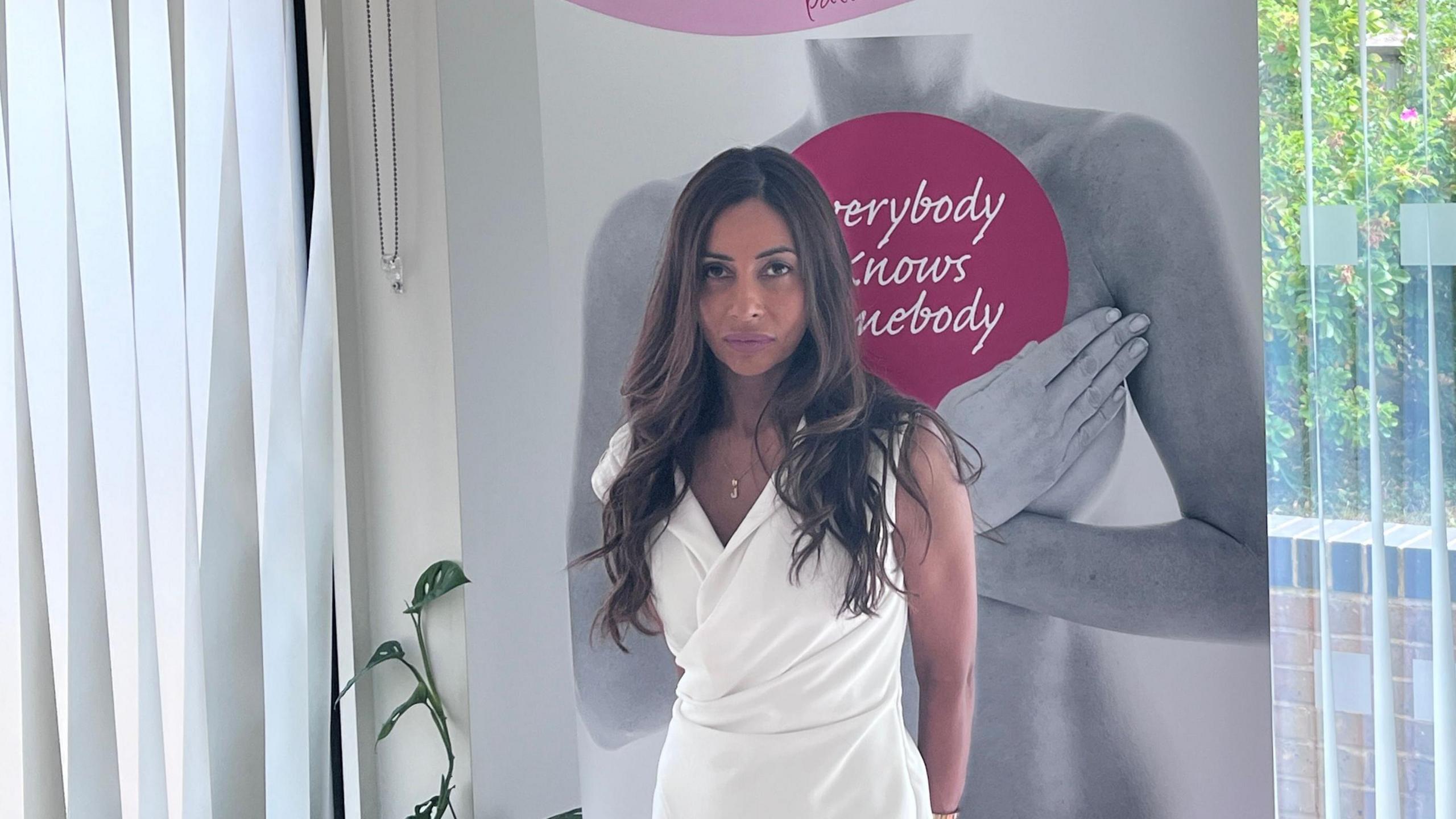 Jothie Walford pictured standing in front of a pink and grey sign that says Worcestershire breast unit haven. She is wearing a white dress and has long curly brown hair.