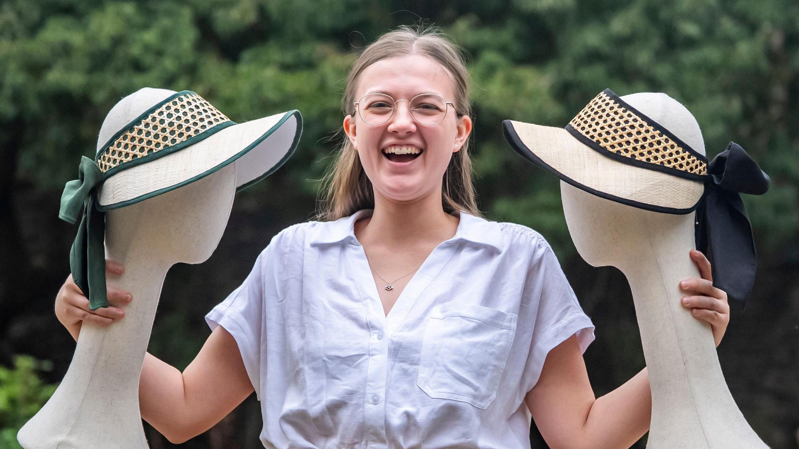 A young woman wearing a white blouse and glasses smiles as she holds up the heads of two mannequins, both of which are wearing cream-coloured hats which she designed.
