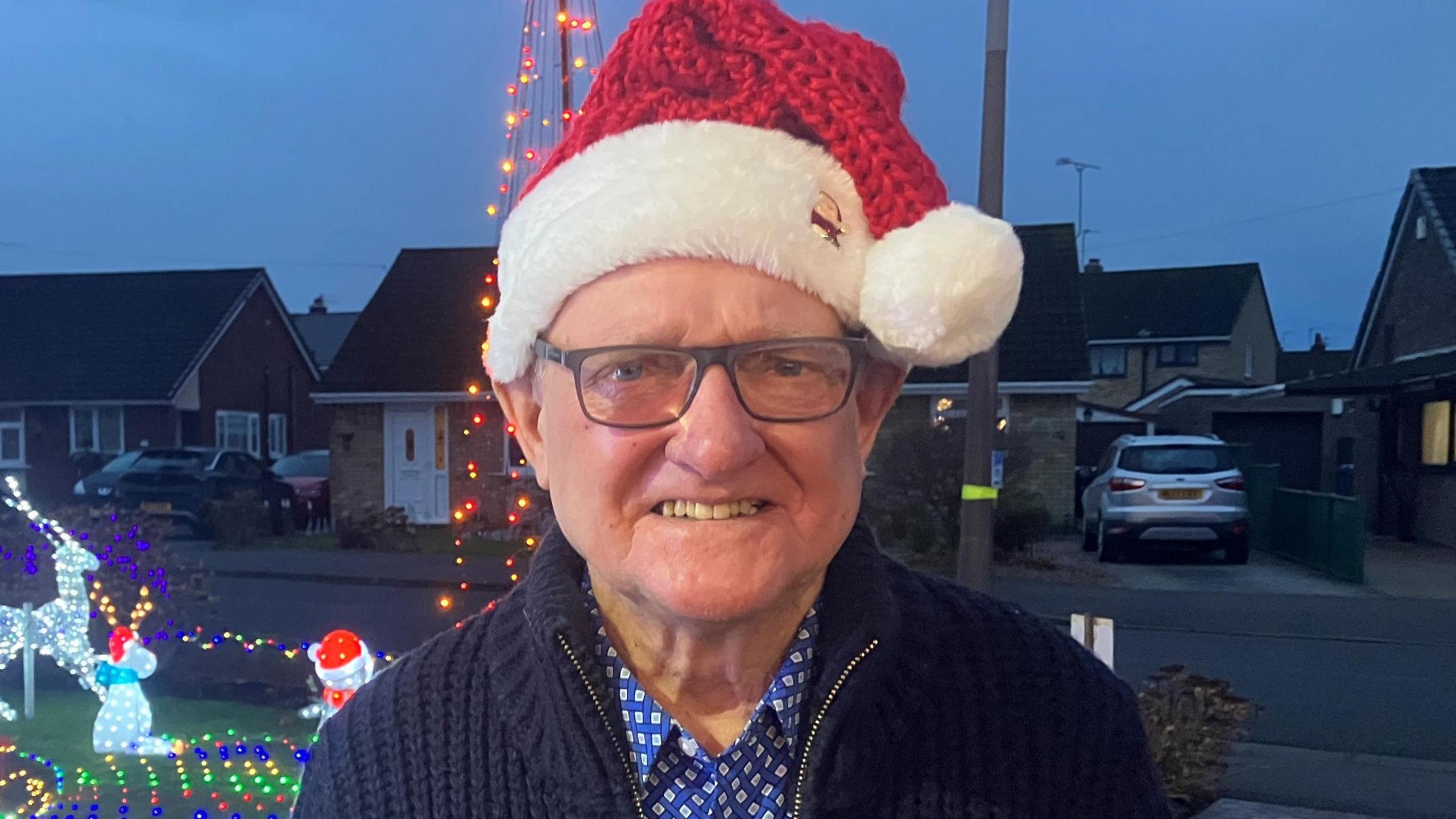 Bryan McAndrew, who has black glasses, smiles on the drive of his home, which is covered in brightly lit Christmas figures. He is wearing a red-and-white Santa hat.