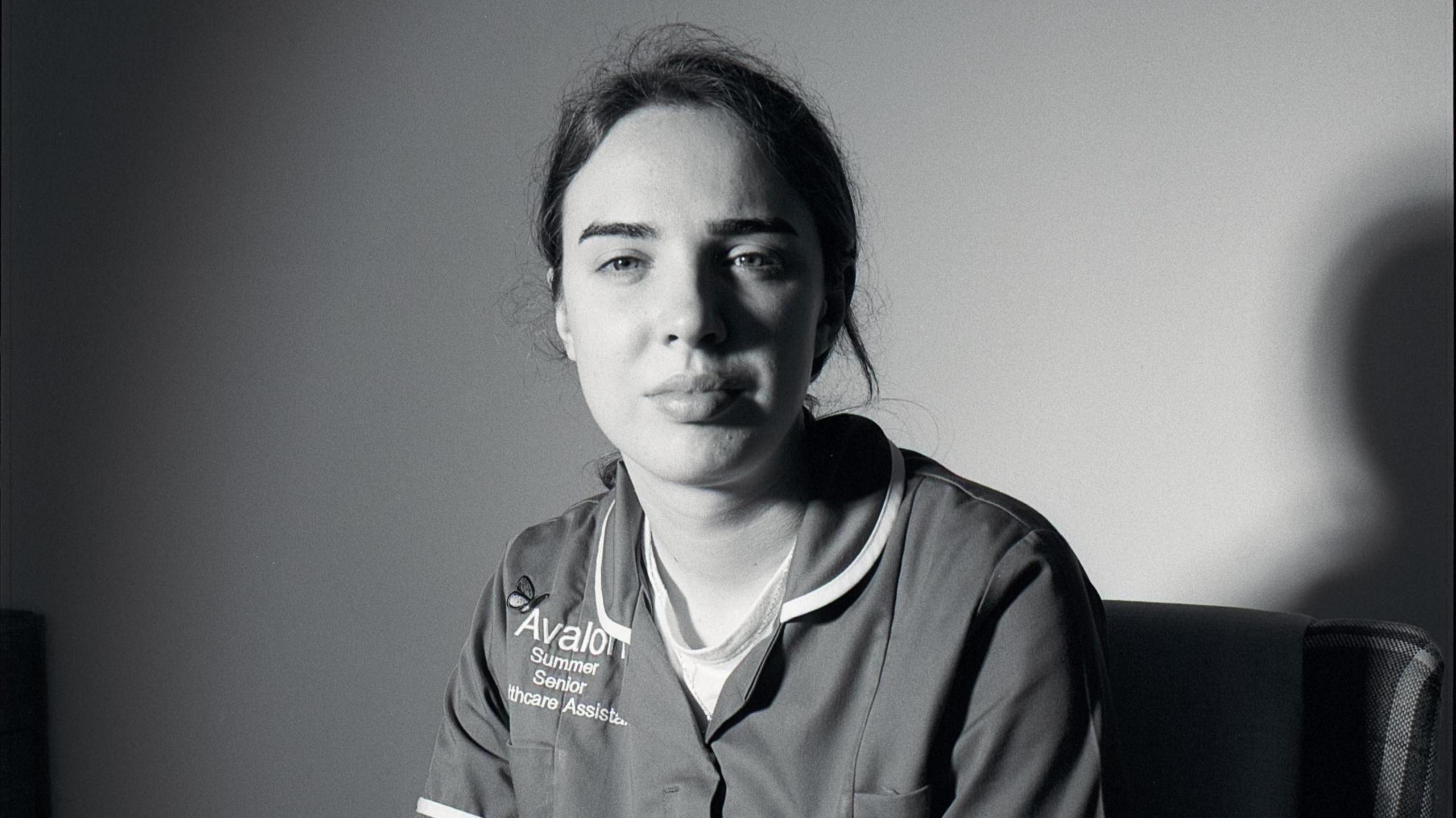 Black and white portrait of Summer Woodley. Her hair is tied up and she is wearing a care uniform with the Avalon logo on the chest. She is looking directly at the camera. 