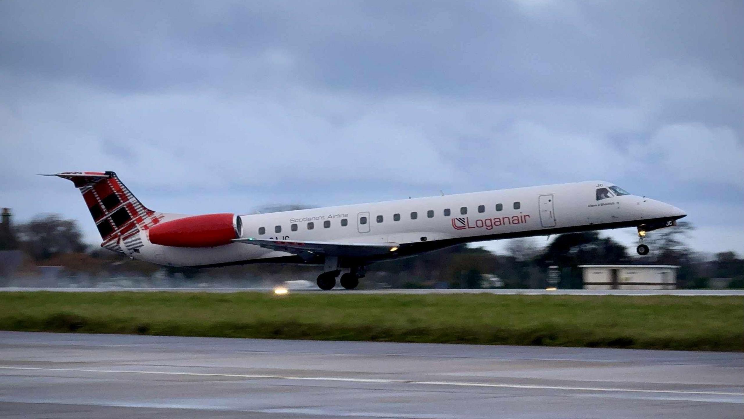 A Loganair plane takes off. Smoke from the rear tires can be seen. The front of the plane is in the air while the rear wheels are still grounded. The plane has a red tartan pattern on its fin. It is a cloudy day.