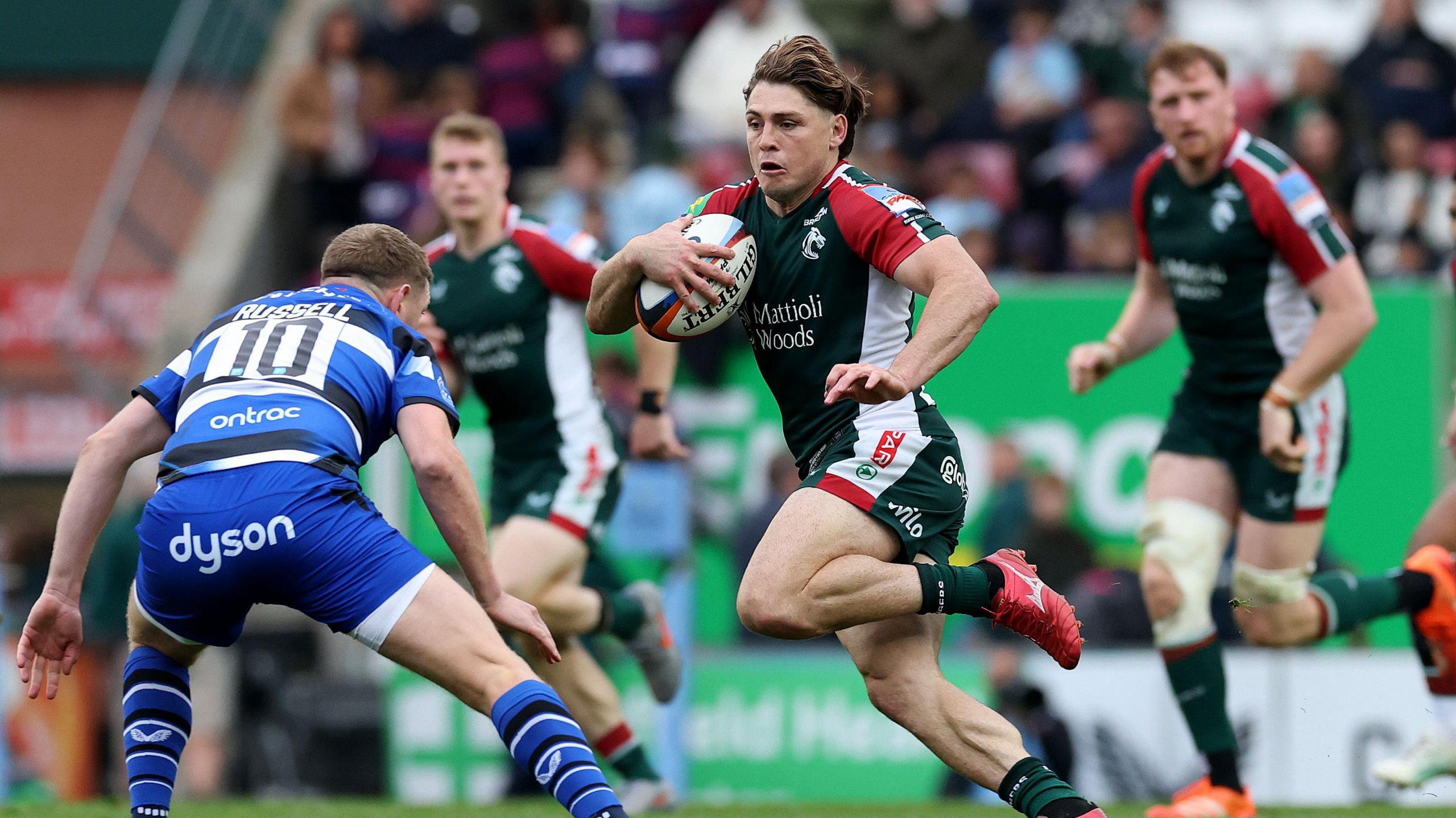 James O'Connor wearing the Leicester bottle green strip runs with the ball in hand as Finn Russell, wearing the blue, black and white of Bath prepares to tackle him. Two other Leicester players look on in the background.