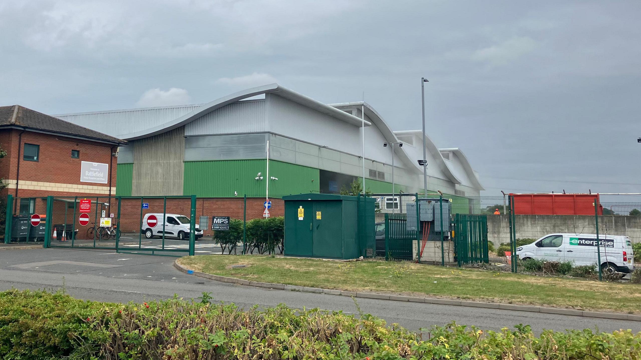 A general view from the road of a large white, green and brown industrial building behind a green fence. Vehicles are parked inside the fence and white smoke can be seen coming from the premises.