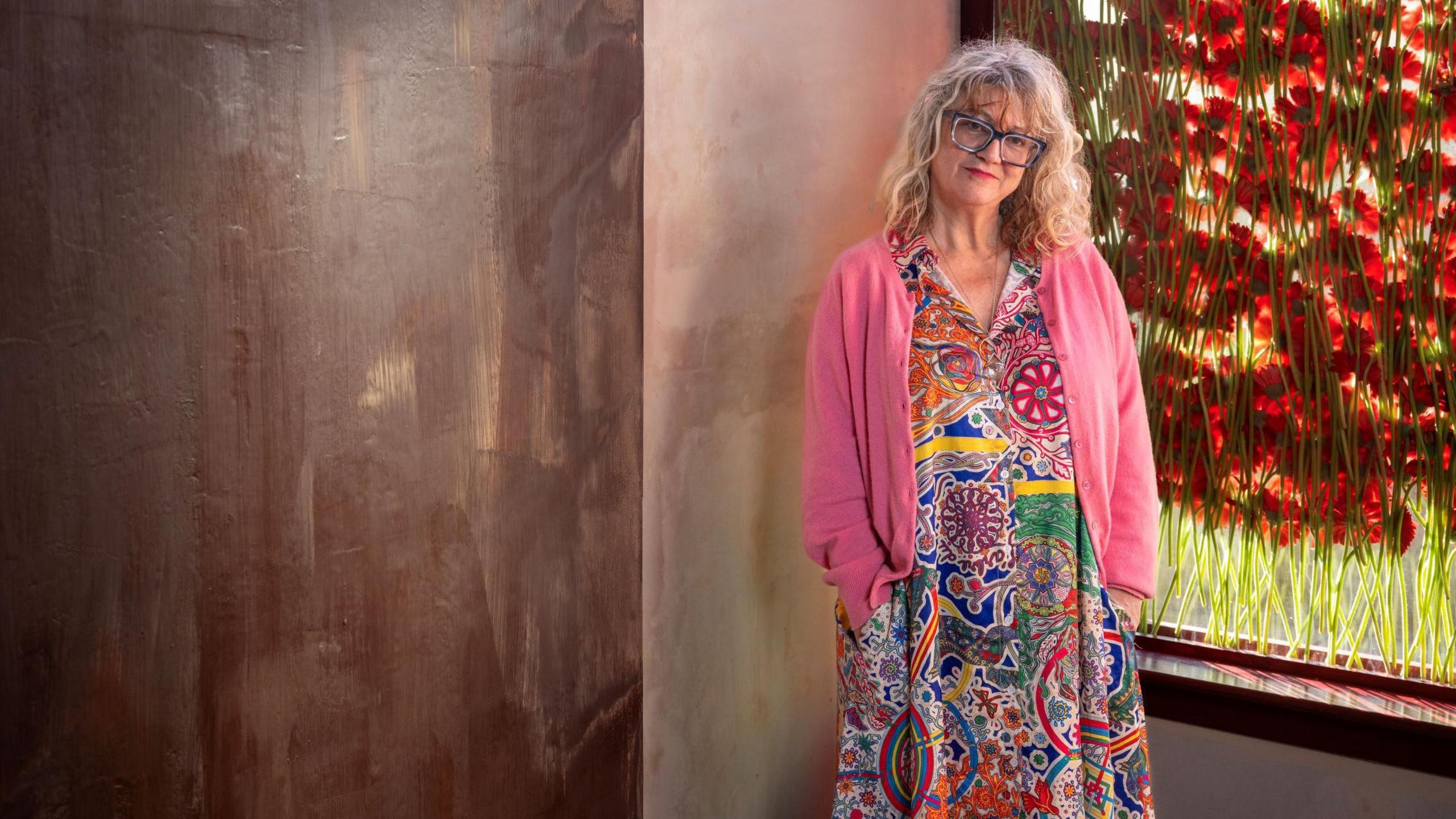 Artist Anya Gallaccio stands at the window in her art installation, beside a wall of chocolate. There are red poppies on the window behind her. Anya is wearing a floral patterned dress and pink cardigan