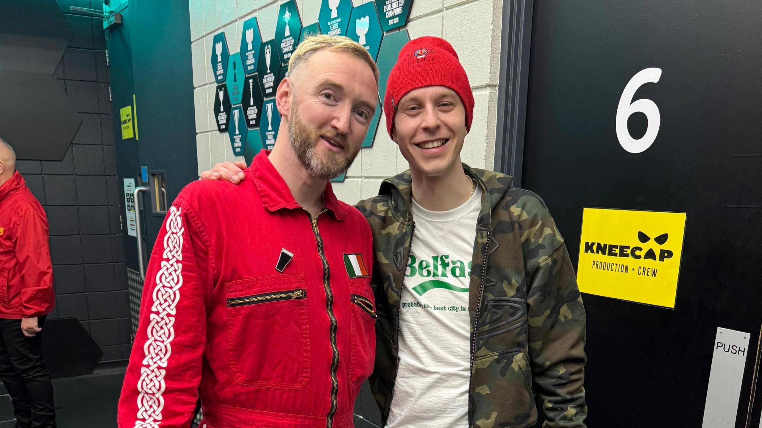 Two men stand in front of a black door which has a yellow sign that reads "Kneecap. Production + Crew." The man on the right has on a camo jacket with a white T-shirt underneath. The man on the left has blonde hair and a red/brown beard. He wears a red boiler suit with three golden zips. There is an Irish badge on the left side of his chest.