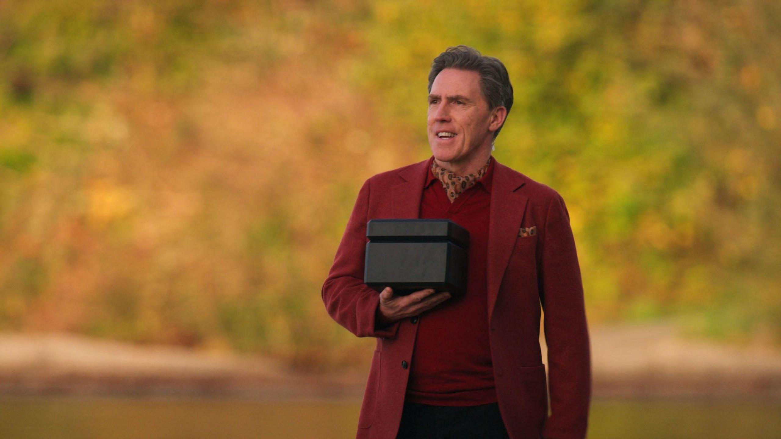 Rob Brydon. He is holding a black box and has a burgundy suit on with a leopard print neck cravat. There are bushes blurred in the background.