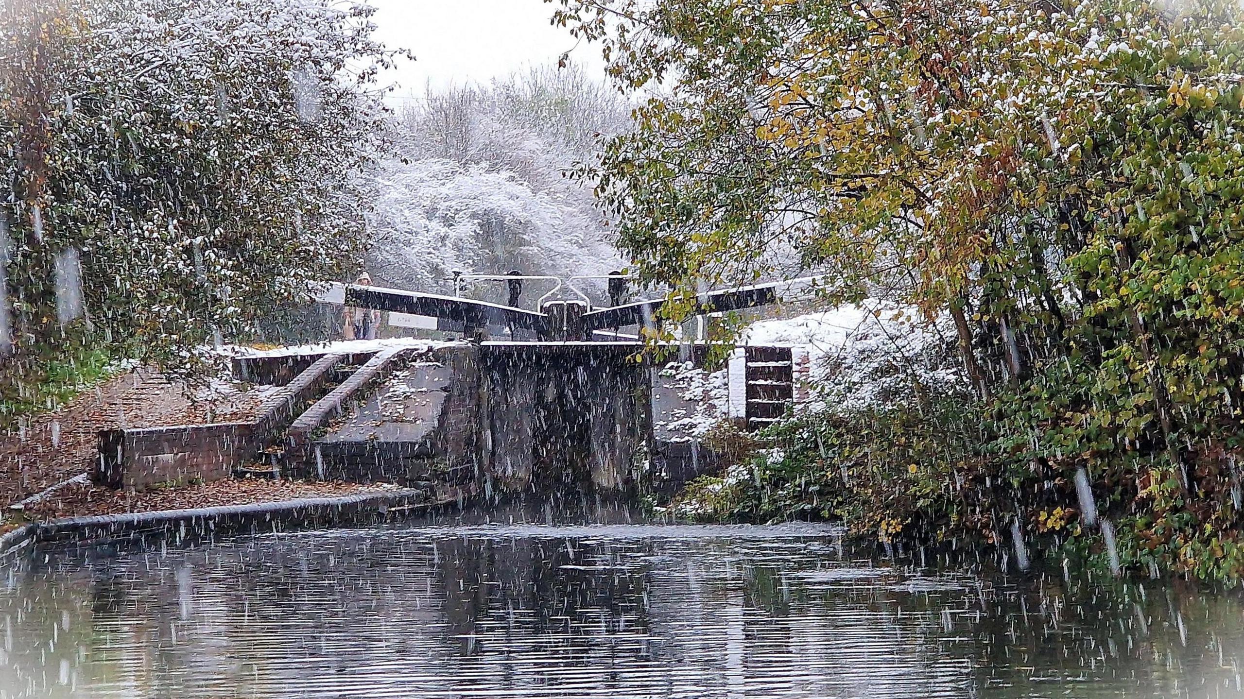 Trees sit on either side of the canal with a canal lock at the top of the water. In the background are trees covered in snow.