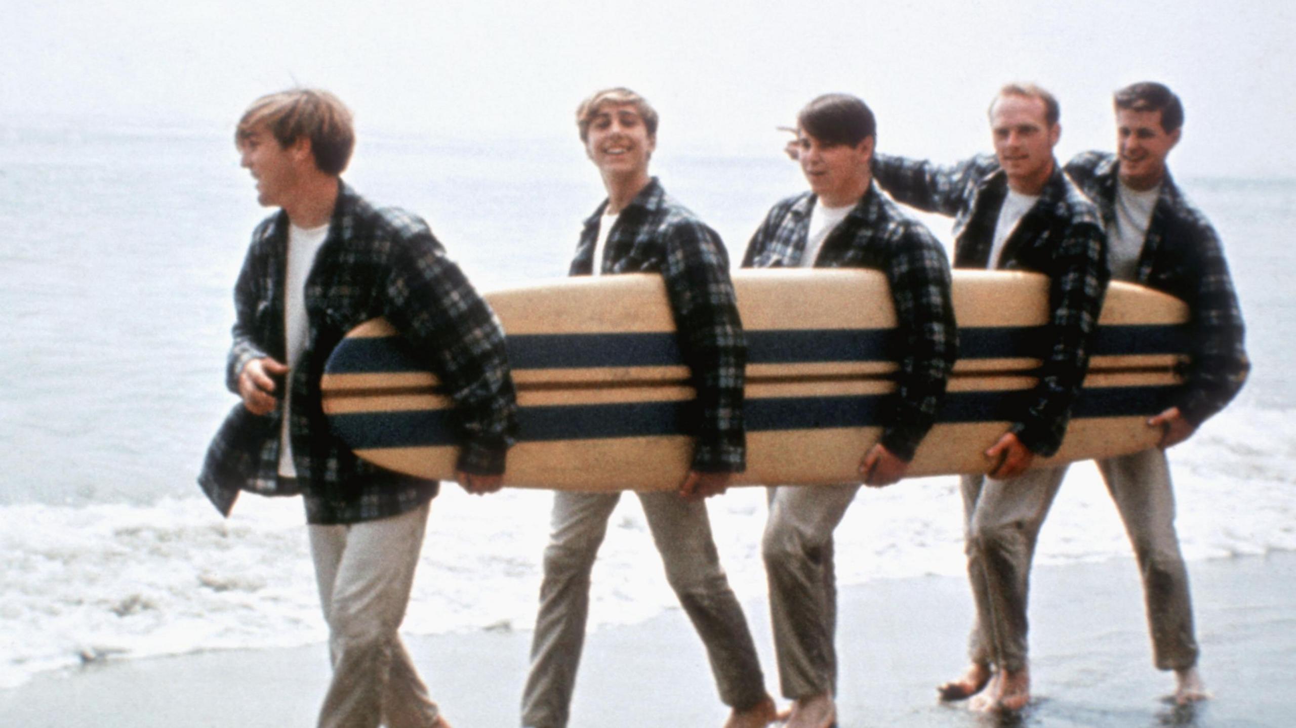 The Beach Boys carry a surfboard between them and walk along the beach in matching plaid shirts and light brown trousers.