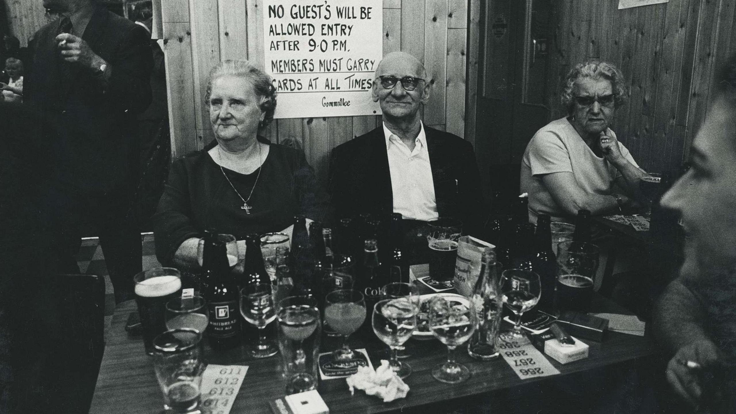 A black and white image of an elderly man and woman sitting at a table in a pub - the table in front of them has many glasses on it. 