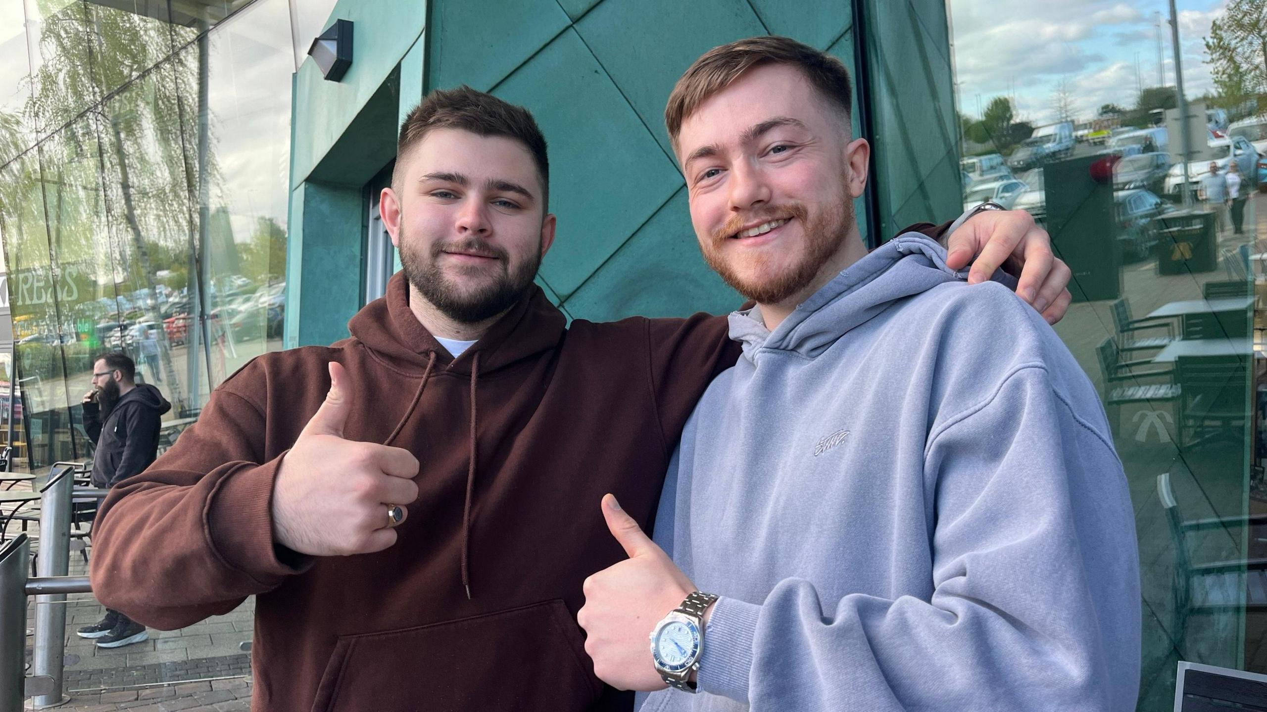 A man in a brown hoodie stood to the left of a man in a lilac hoodie. Both men have one arm around each other and are holding their thumbs up.