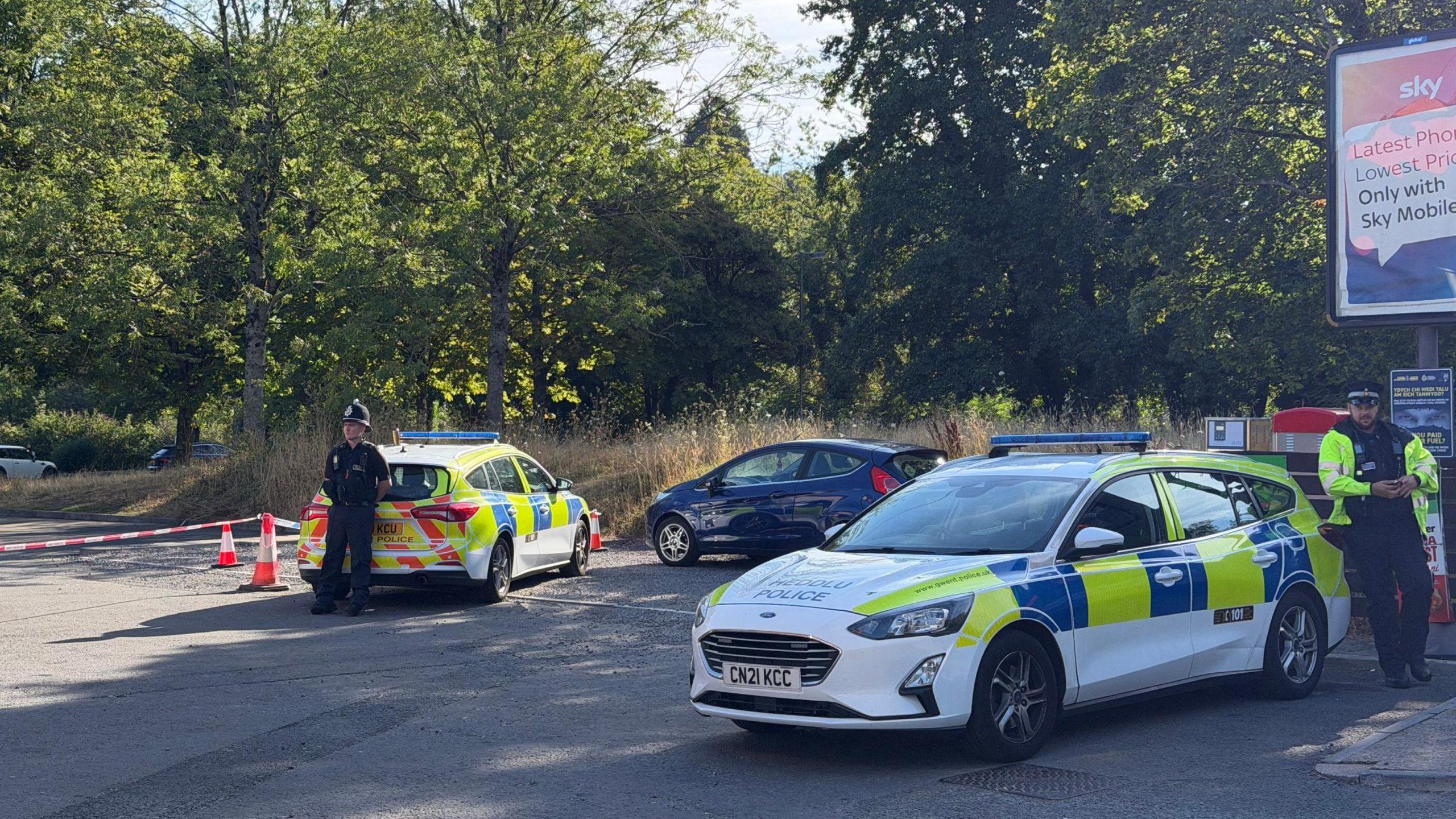 A police cordon with two police cars and a police officer.