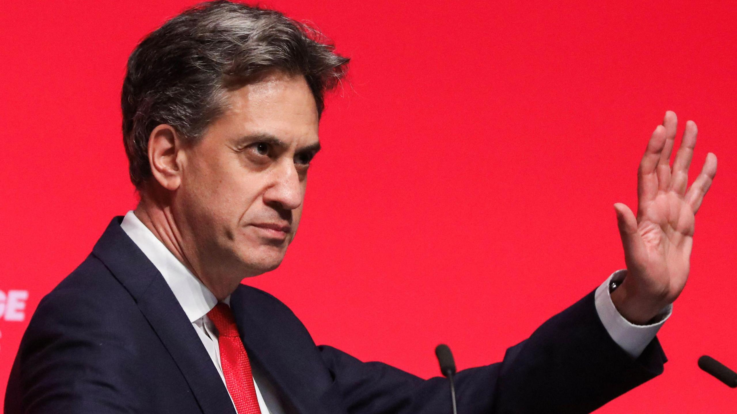 Ed Miliband raising his hand while giving a speech with a red backdrop