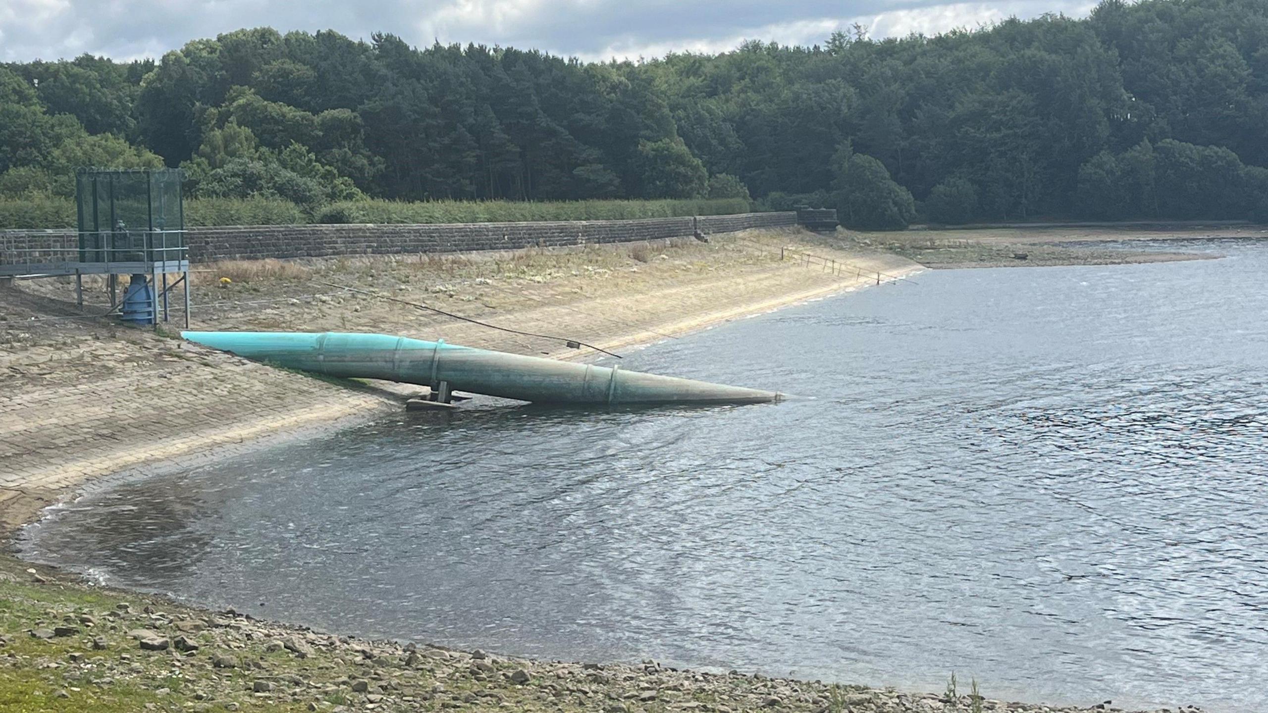 A dried out reservoir in Leeds. A large turquoise pipe can be seen going into the water. There are trees in the background.