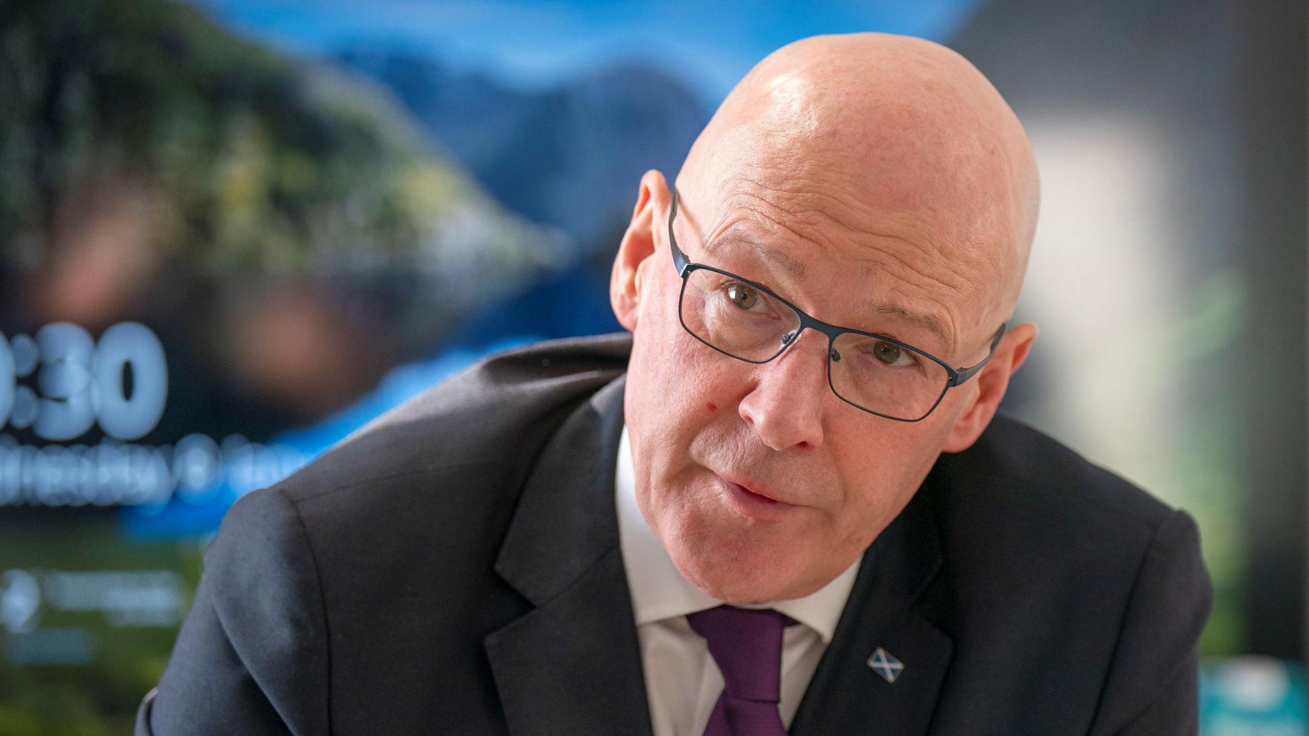 John Swinney, in mid conversation with someone off camera - he appears to be listening to them. He is bald, with glasses and and dark suit with purple tie and a lapel pin with a Scottish flag on it.  