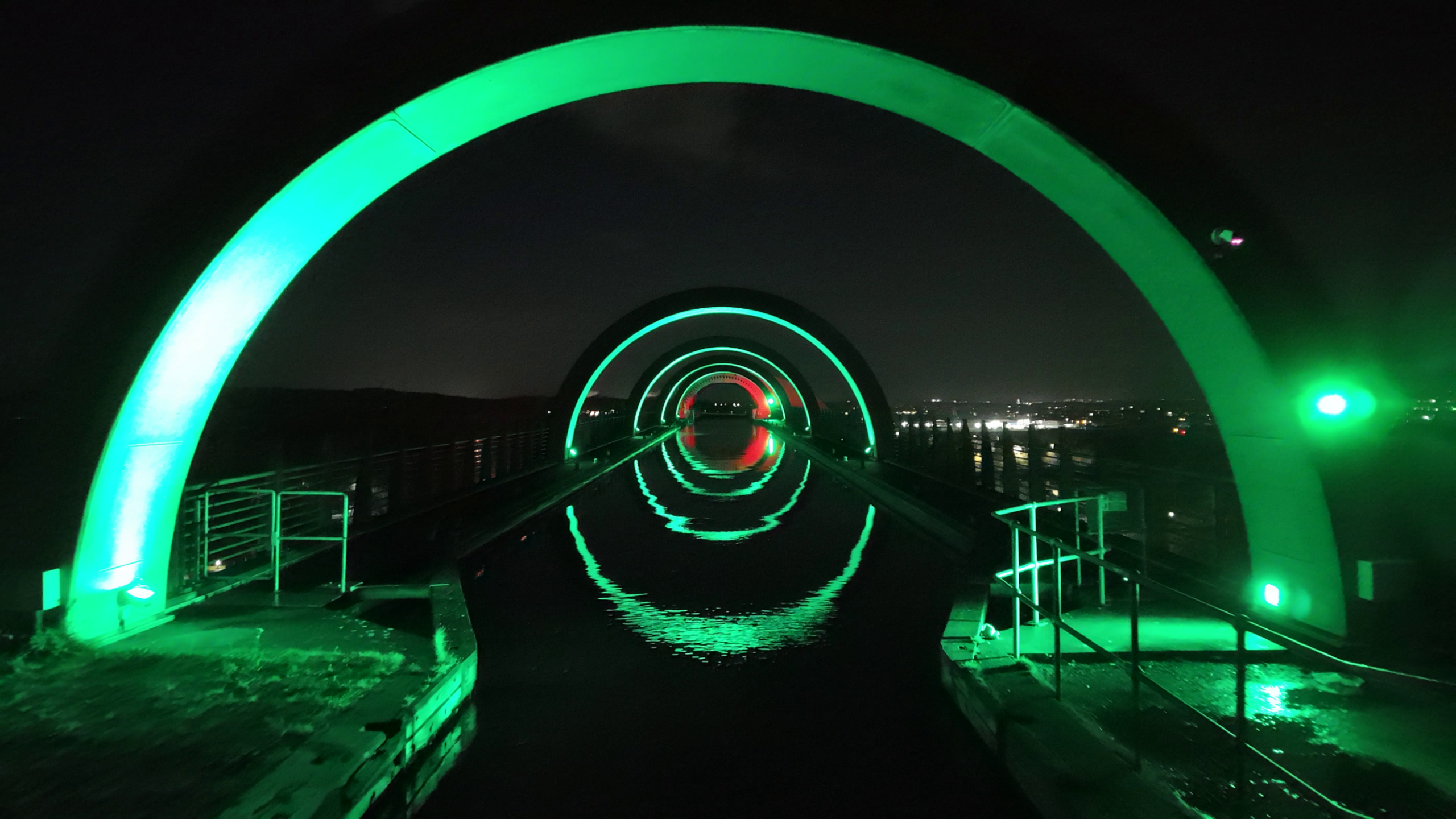 A series of illuminated green arches create a mesmerising tunnel effect over a calm waterway, their reflections forming perfect circles. The structure is dramatically lit against the dark night sky, with distant town lights faintly visible in the background. Metal railings and platforms frame the foreground.