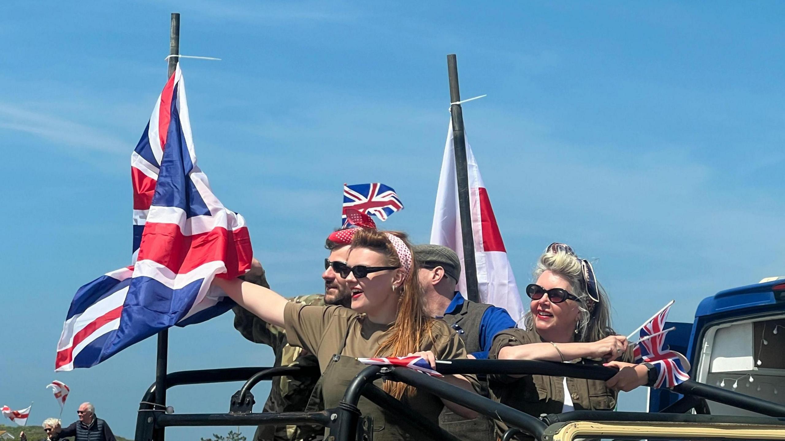 People in vintage military colours waving the Union flag as they stand on top of a Land Rover