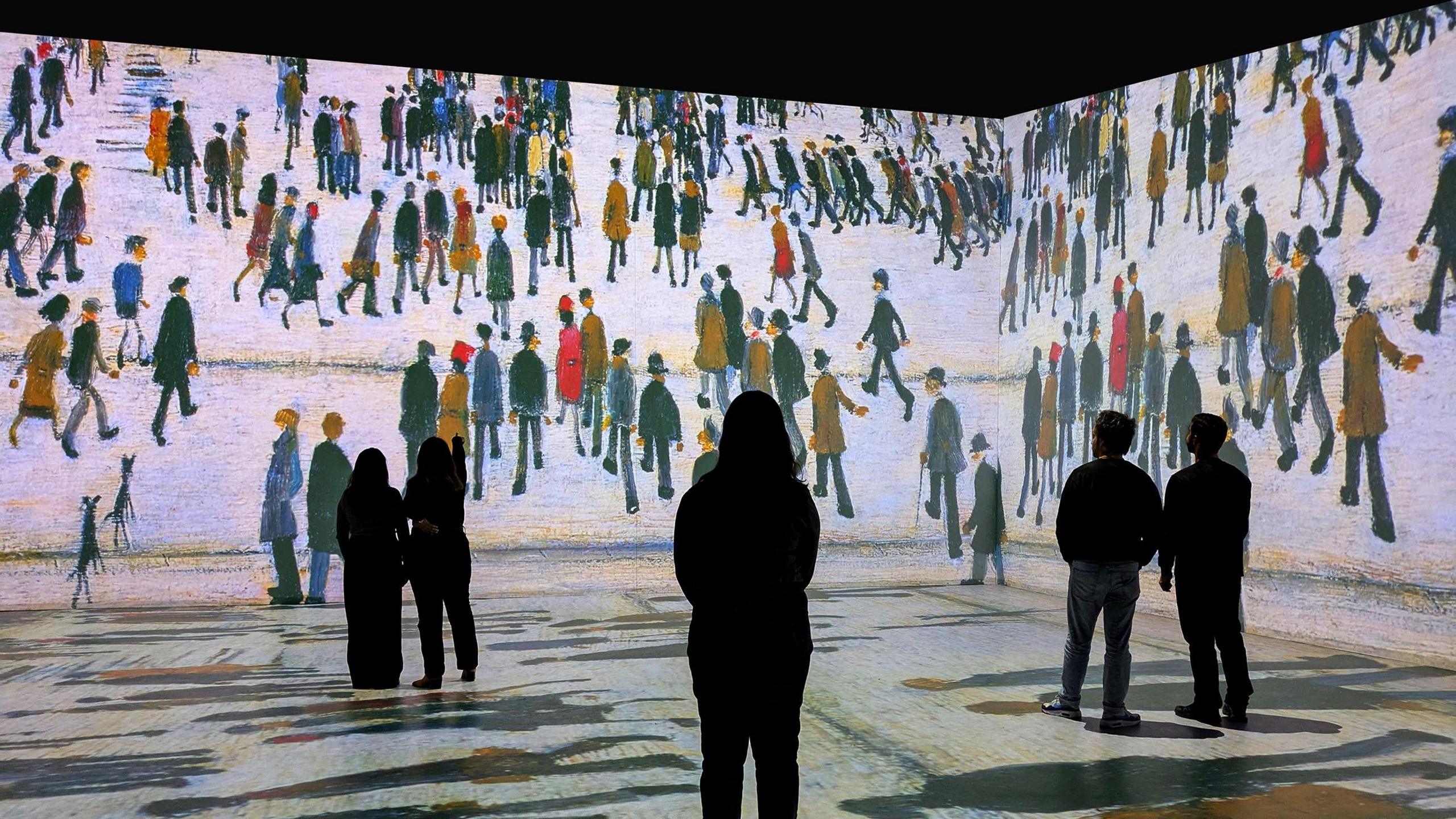 A woman stands in the middle of a 3D-display, featuring a LS Lowry painting superimposed on the floor and walls of a room in an art gallery. She is stood in front of groups of other people with their backs turned away from the camera, watching the display.