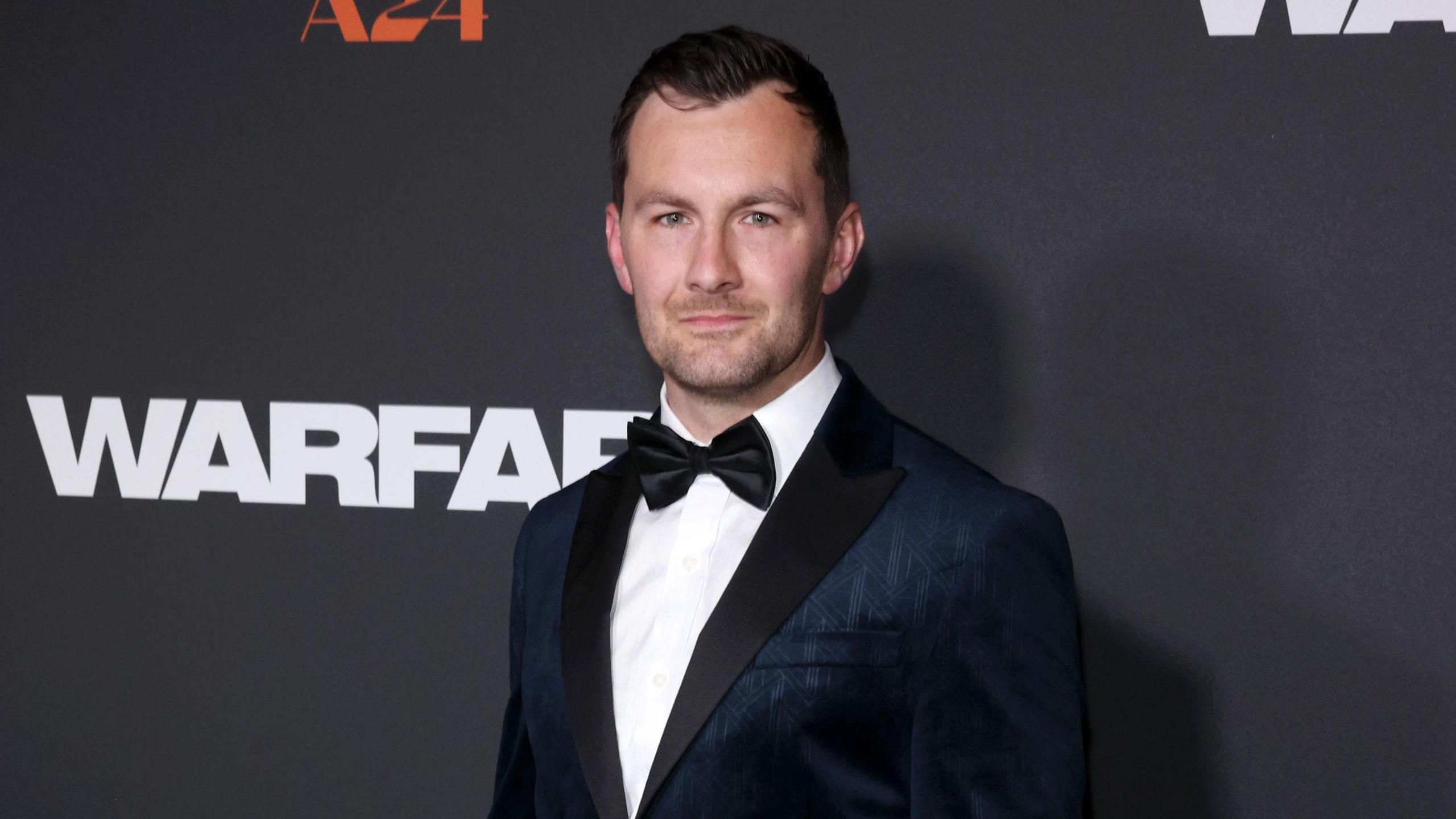 Aaron Mackenzie in a navy tuxedo stands next to a Warfare branded wall, posing for photos at a film premiere.