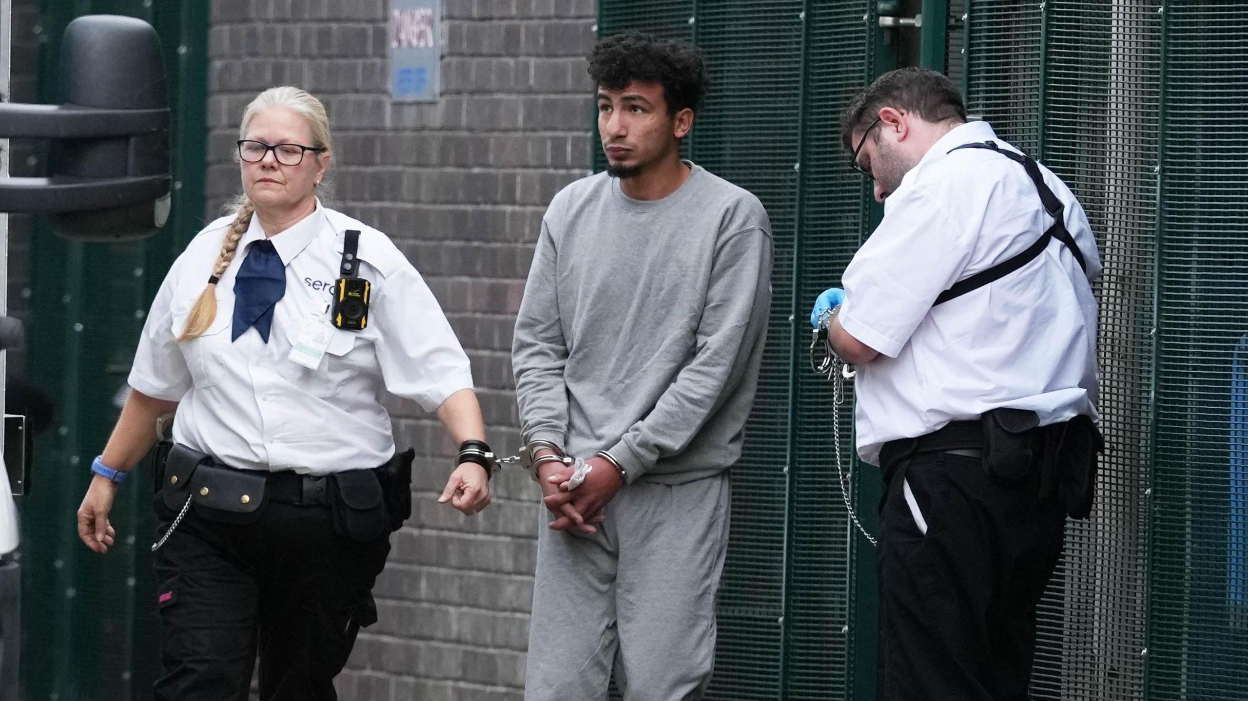 A man in grey clothing and handcuffs is with two security officers beside a brick wall and metal fencing.