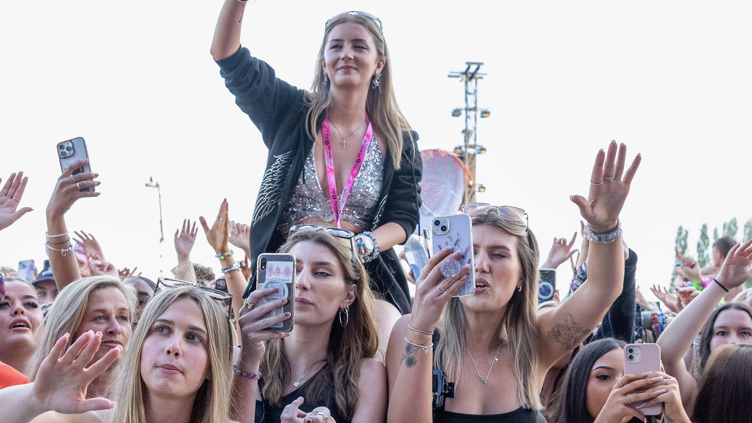 A crowd of mostly young, female festival-goers at Radio 1's Big Weekend. Many are holding phones up, recording a performance. One girl can be seen sitting atop an unseen friend's shoulders, waving her right hand and smiling.