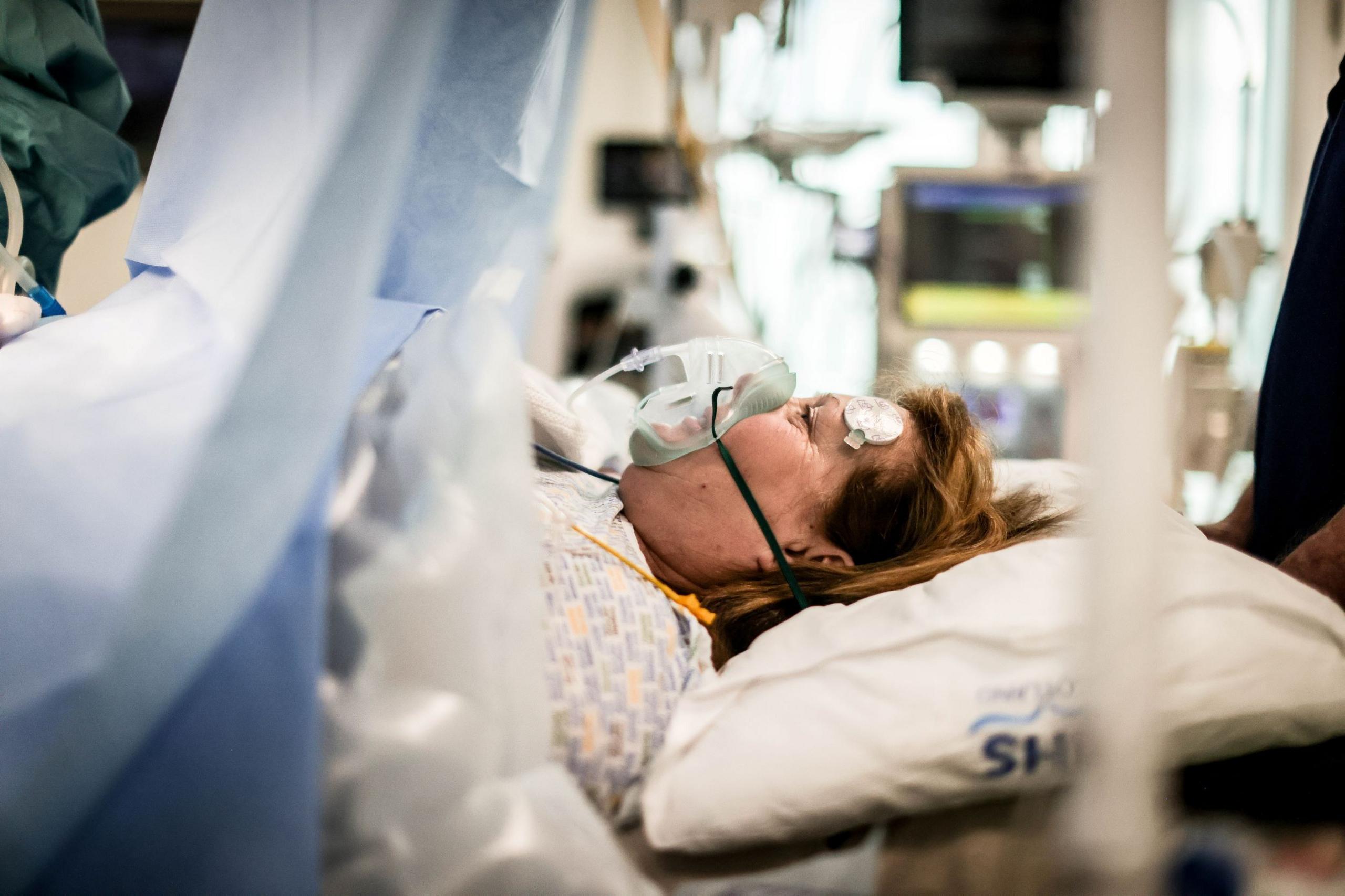 A woman lies on an operating table, in a hospital gown, with monitoring pads on her forehead and an oxygen mask on, with observation monitors out of focus in the rear of the image