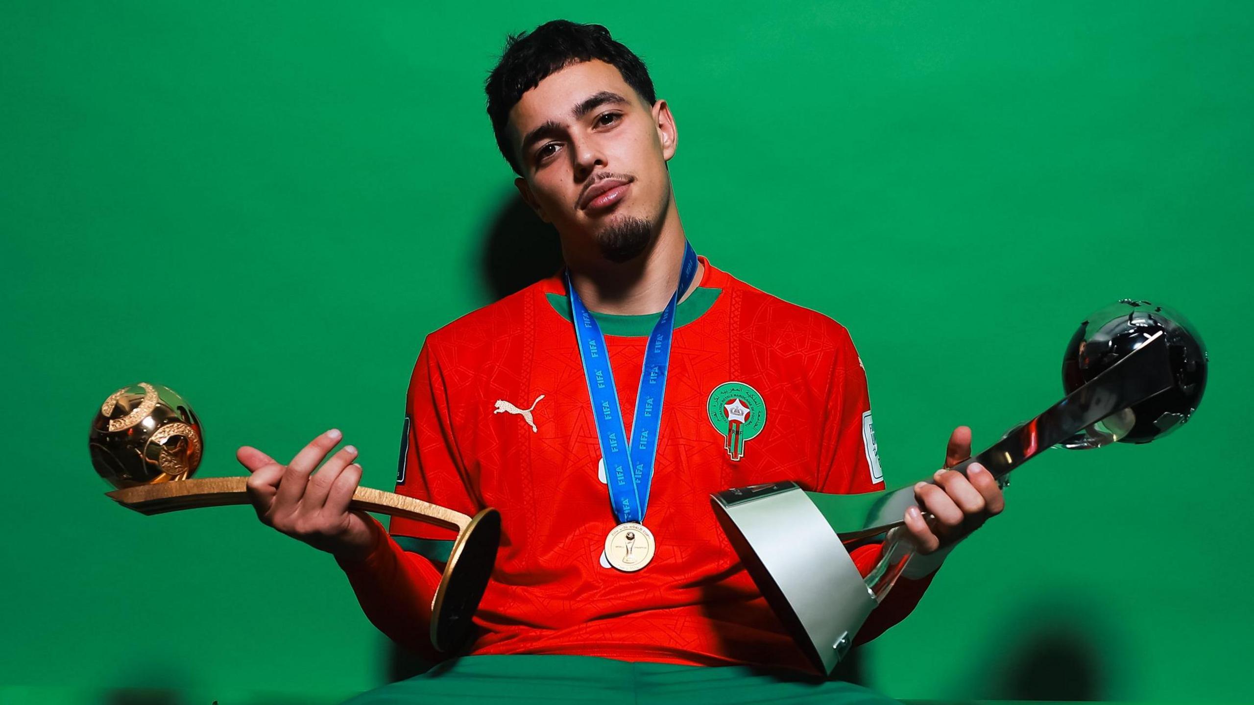 Othmane Maamma, wearing a red Morocco shirt with a gold medal around his neck on a blue ribbon, poses in front of a green screen as he holds the golden ball trophy in his right hand and the Under-20 World Cup trophy in his left hand. He is cocking his head slightly to his right