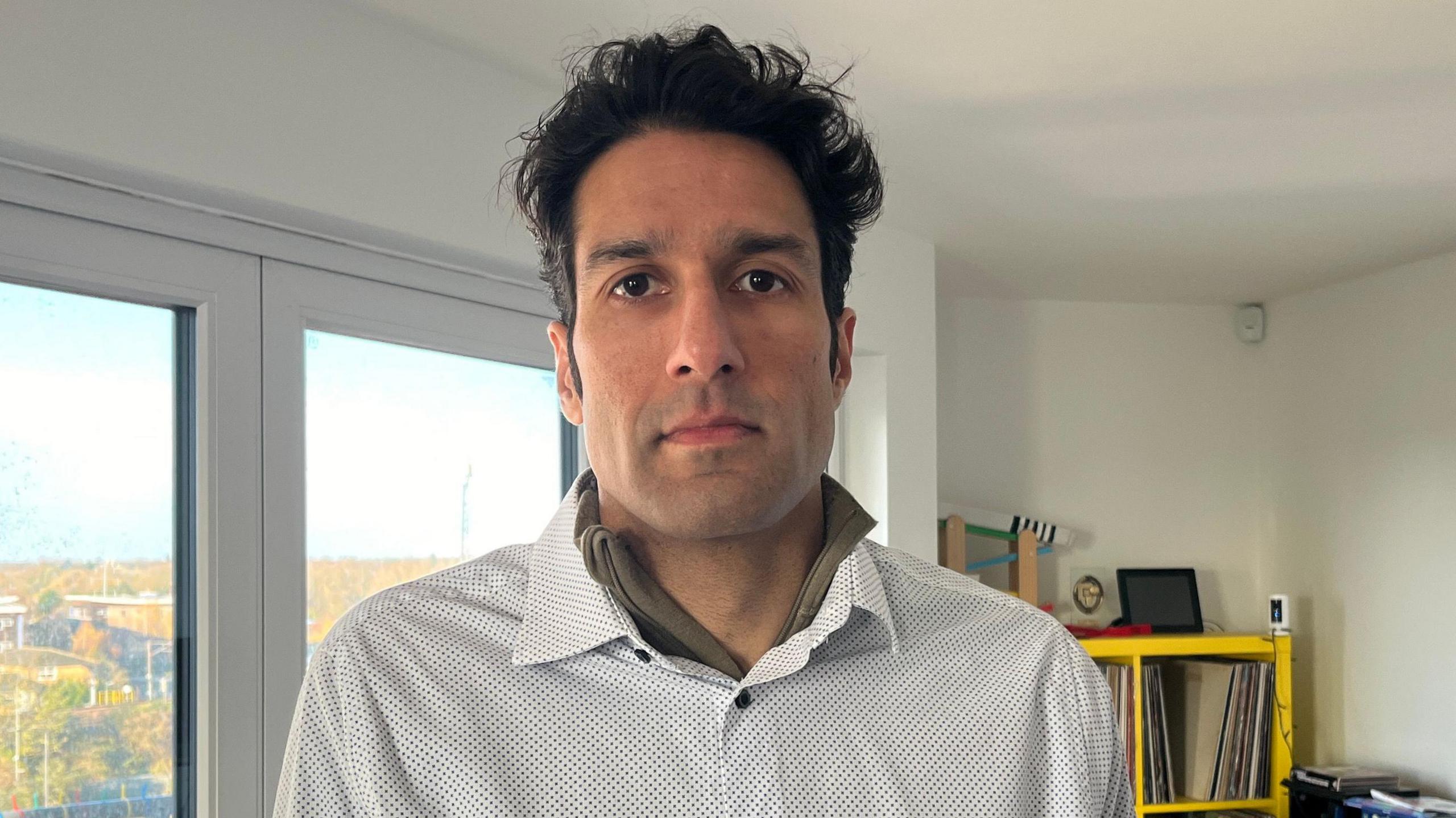 Kason Kalirai inside his flat wearing a white shirt with small black dots on. He has black hair, brown eyes and is not smiling at the camera. 